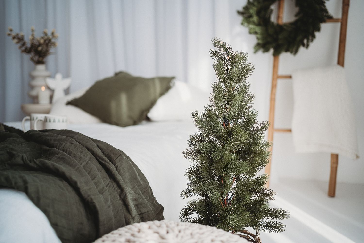 A cozy Christmas bedroom with a small green pine tree, olive pillows, white bedding, and a wreath on a wooden ladder.