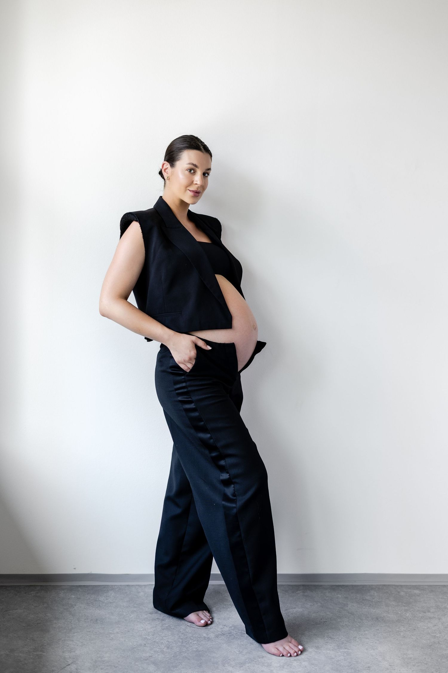 Pregnant woman in black sleeveless blazer and wide-leg trousers, posing against white wall.