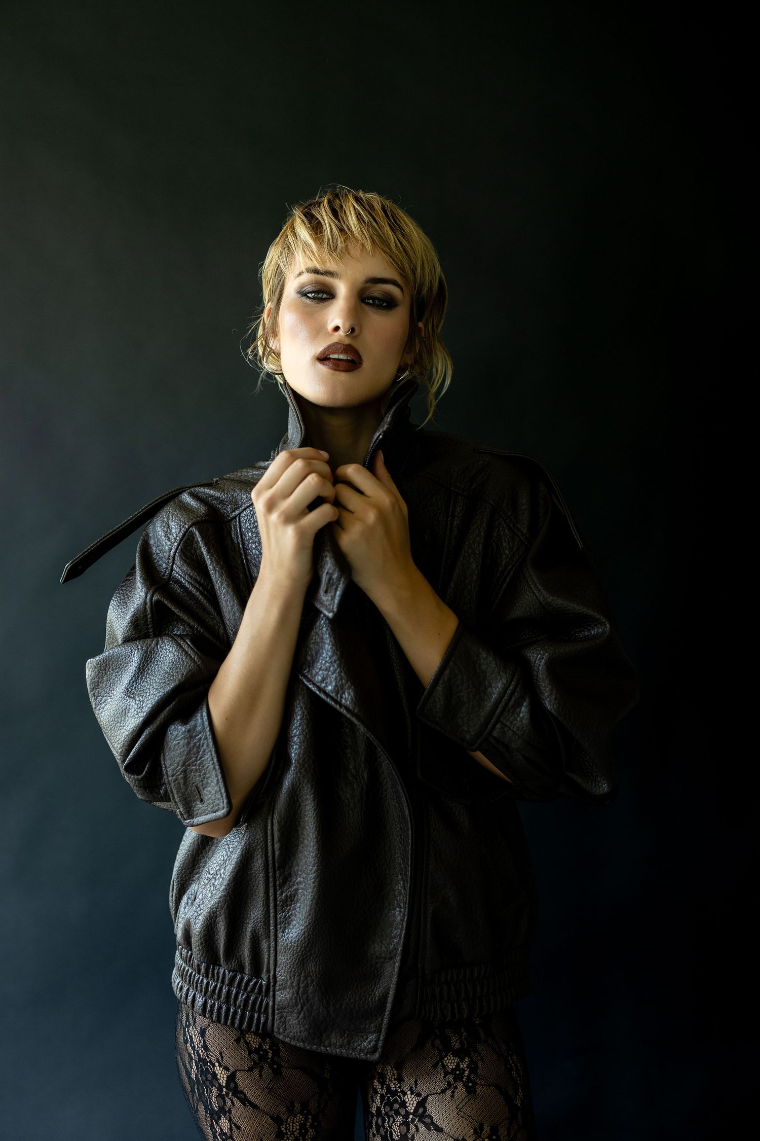 Woman in black leather jacket and lace pants poses dramatically against dark background with bold makeup.