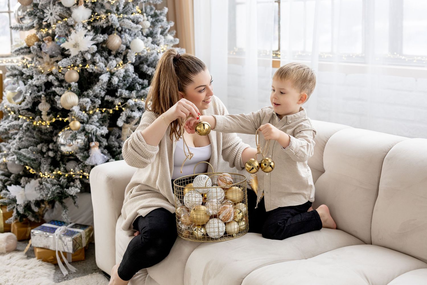 A mother and son in matching beige sweaters share a light moment on a couch near a decorated Christmas tree.