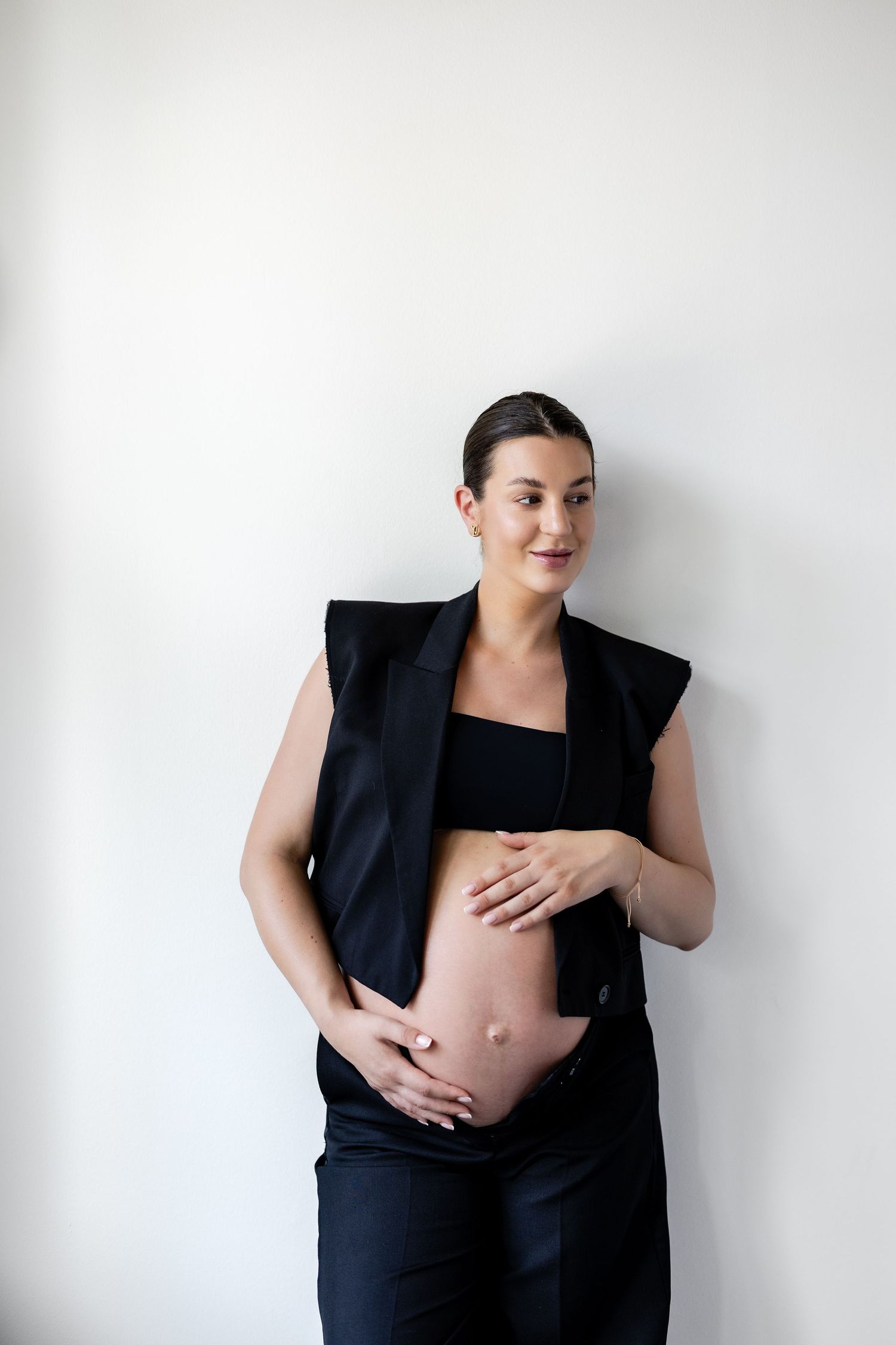 Pregnant woman in black vest and crop top, cradling baby bump against white wall.