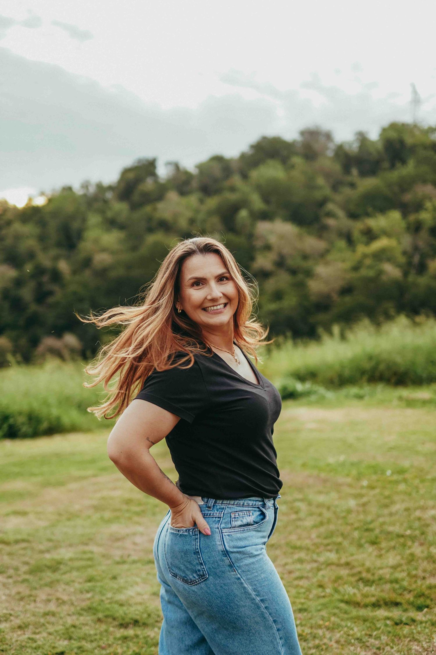 A person in a black top and jeans spins joyfully in a grassy field on a sunny day.