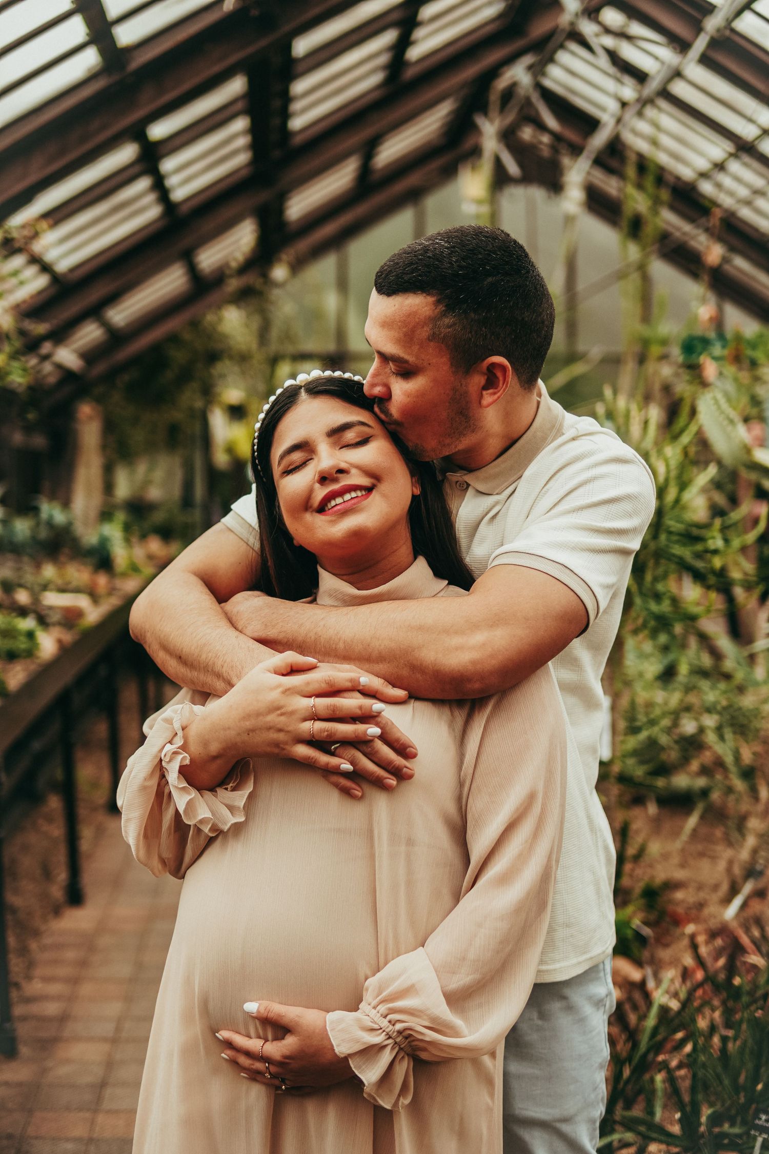 Maternity photoshoot in greenhouse shows tender embrace between expectant couple.