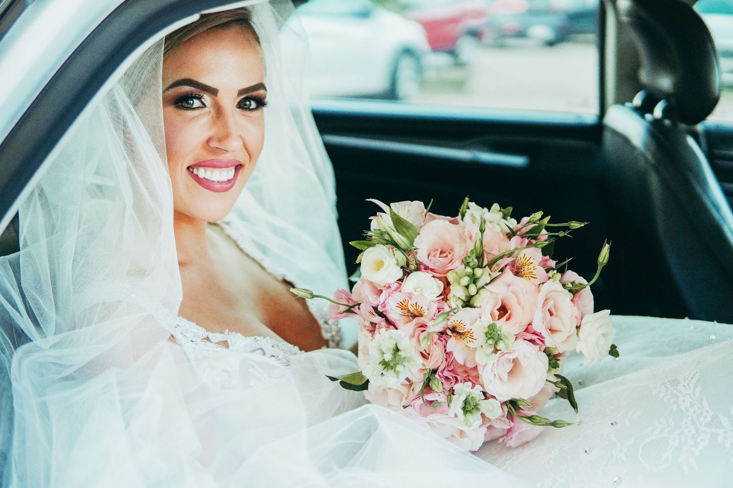 A cheerful bride sitting in a car holding a pink and white wedding bouquet.