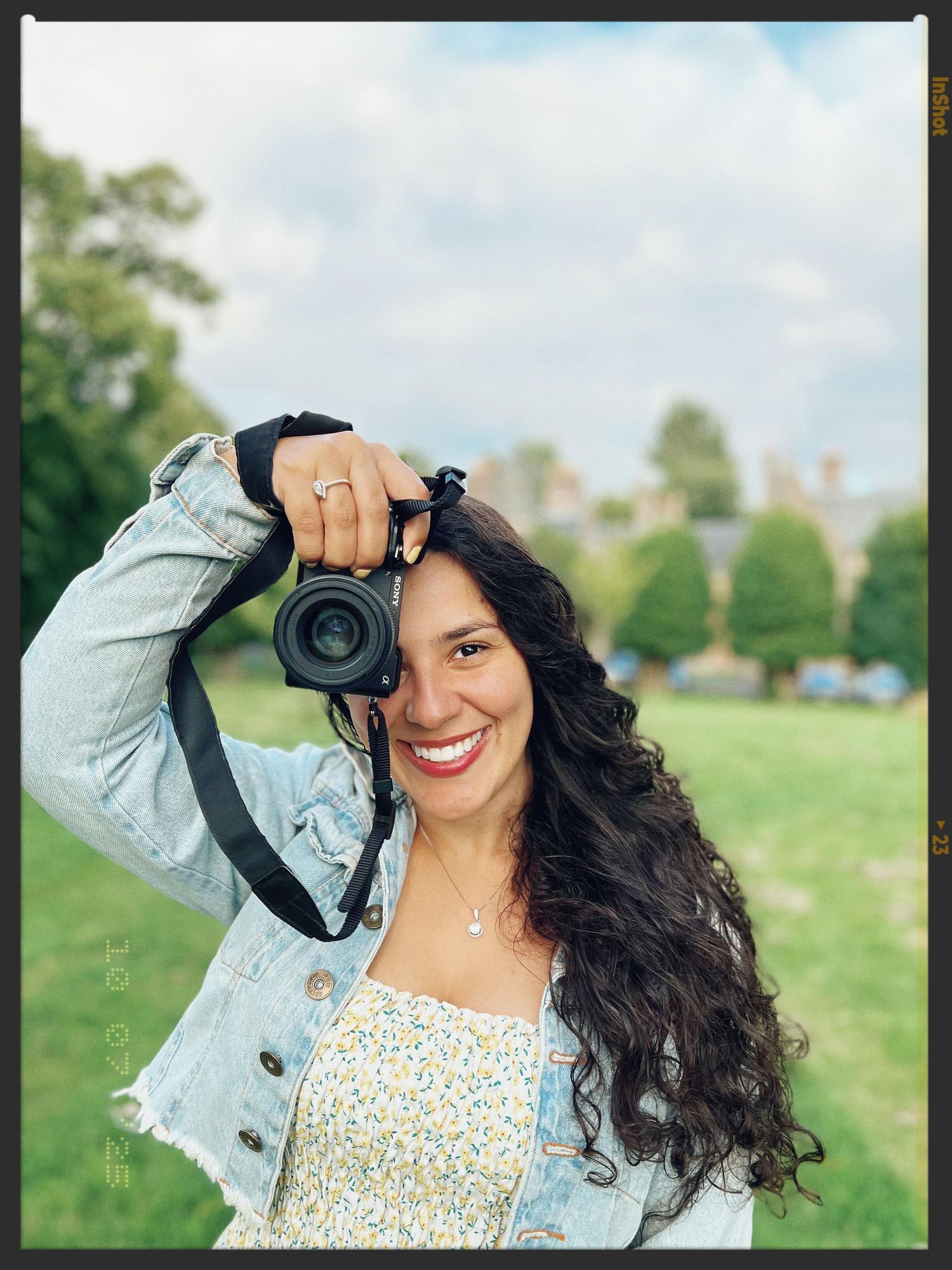 A photographer in a denim jacket smiling while holding a camera in a sunny outdoor field.