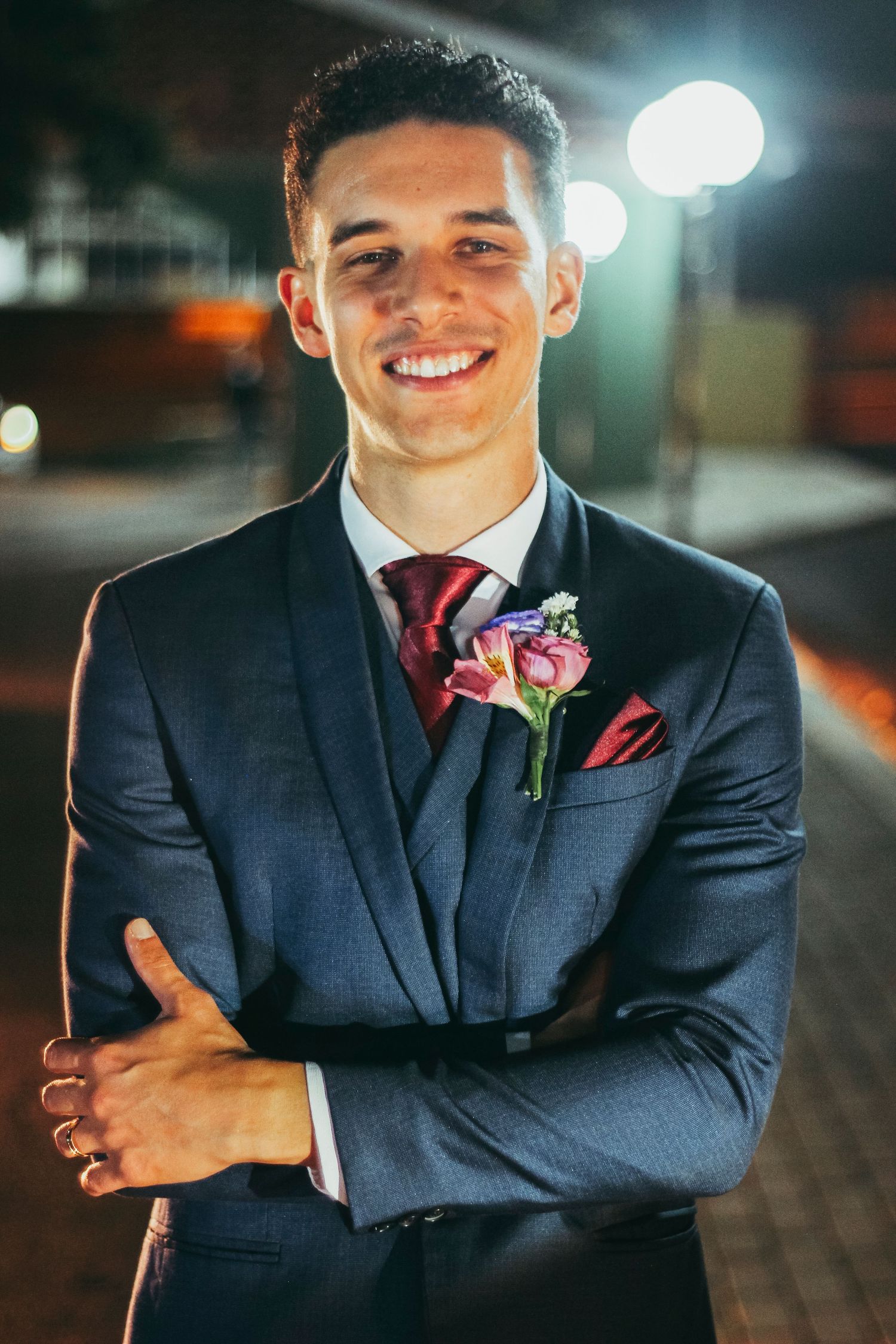 Formally dressed person in navy suit with red tie and boutonniere smiling at night.