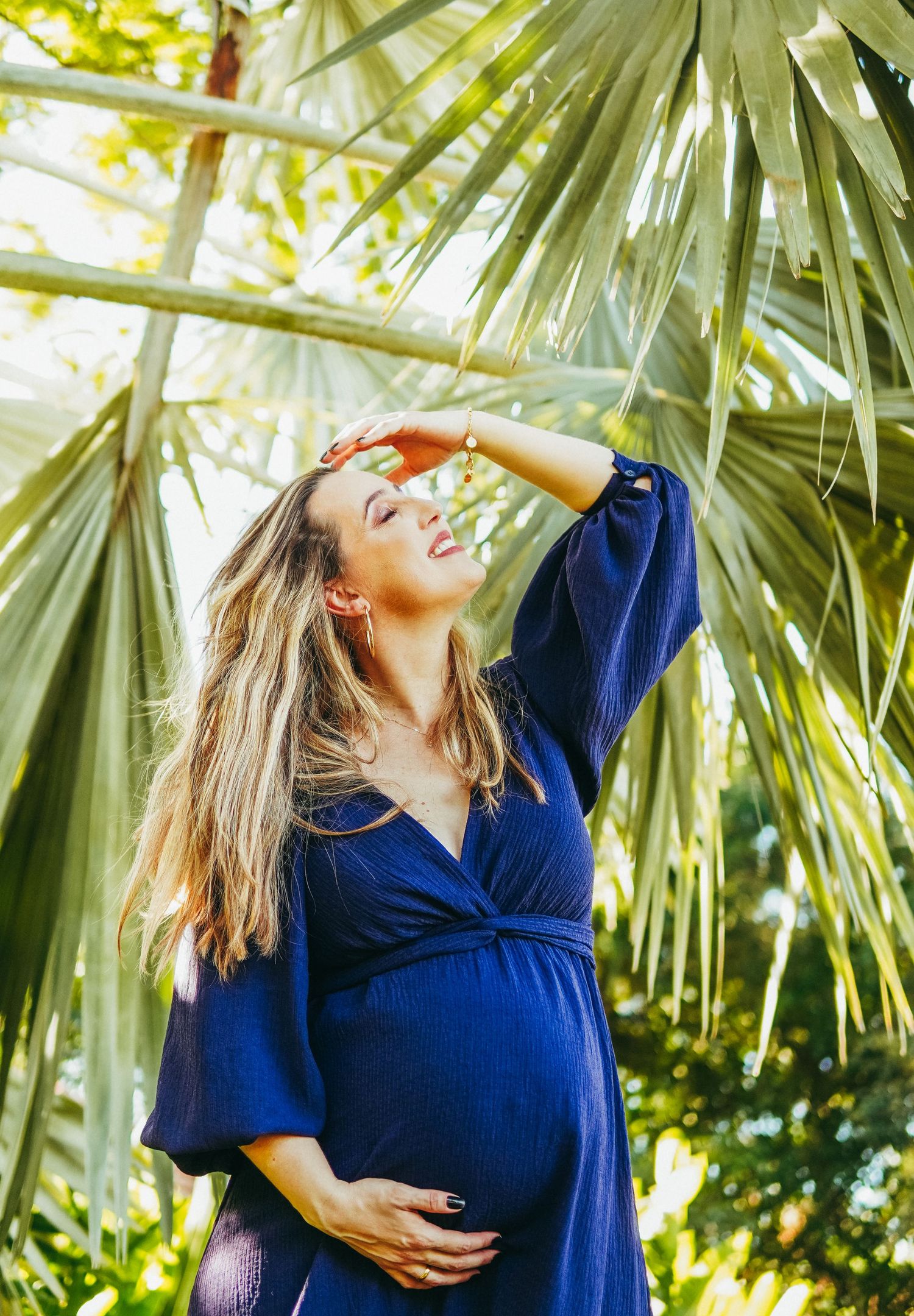 Woman in navy blue dress posing among tropical palm fronds in bright sunlight.