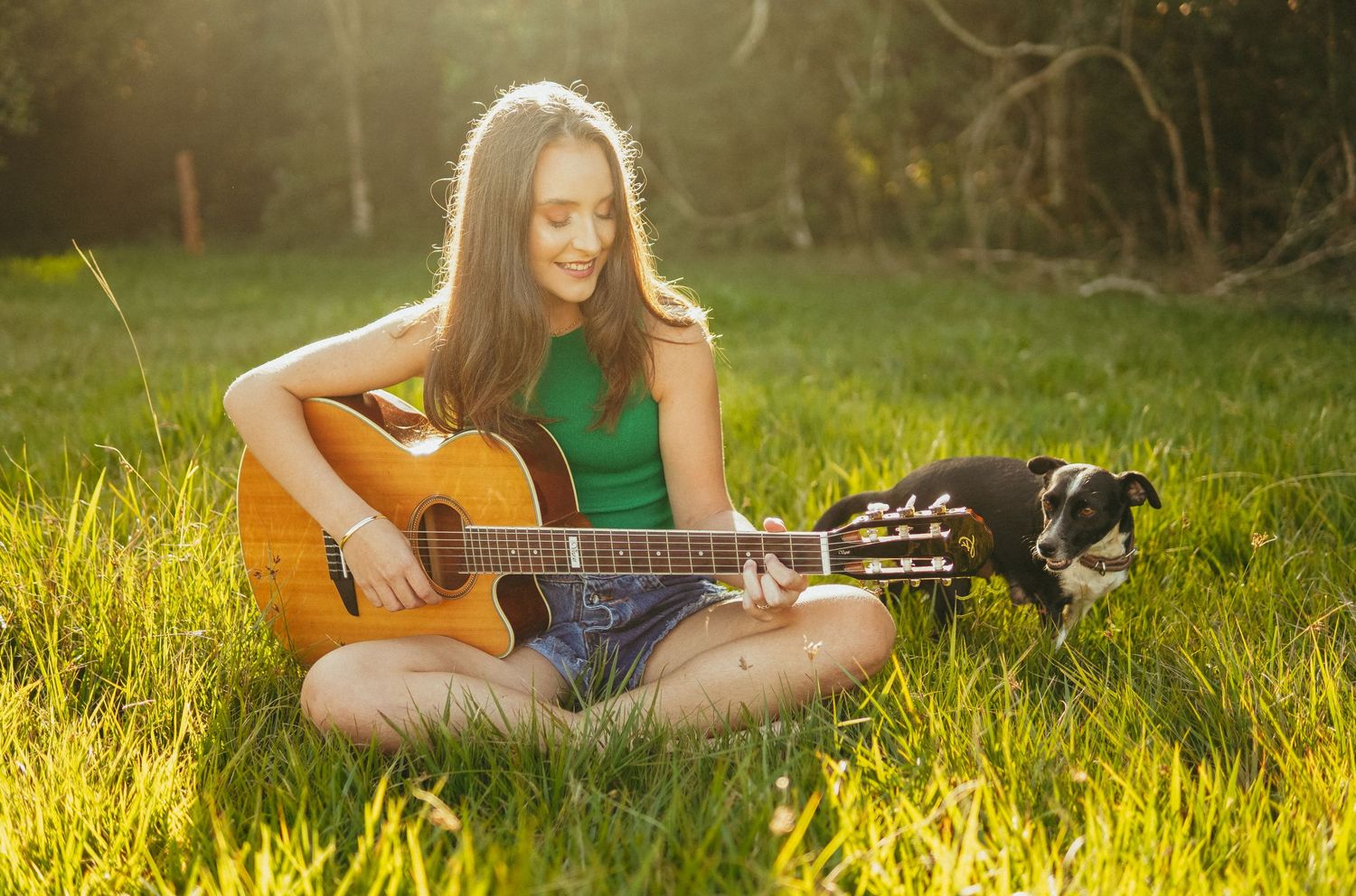 Young woman playing acoustic guitar while sitting on grass in warm sunlight with a black dog beside her.