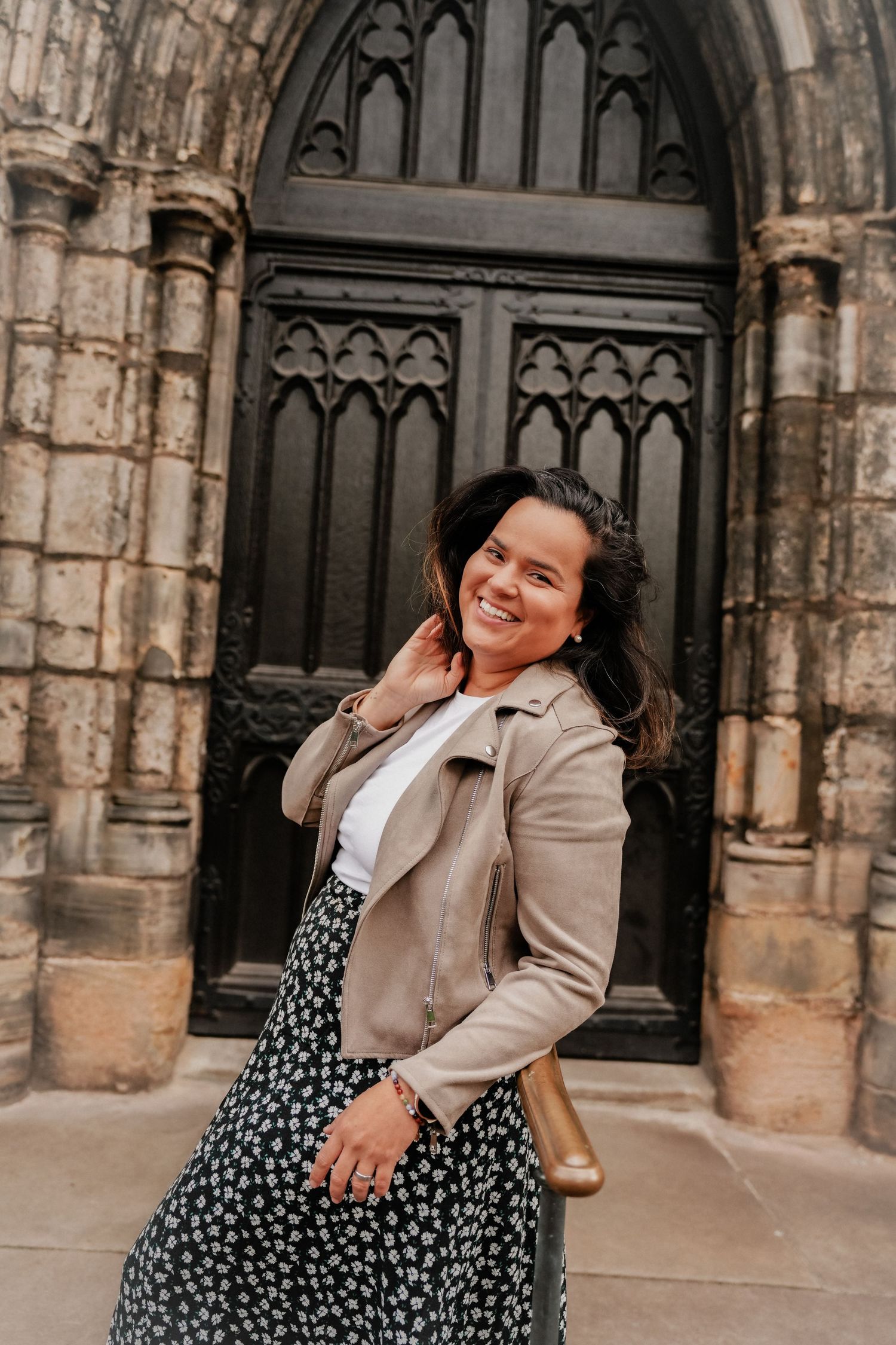 Smiling woman in beige leather jacket and floral skirt poses before ornate Gothic church doorway.