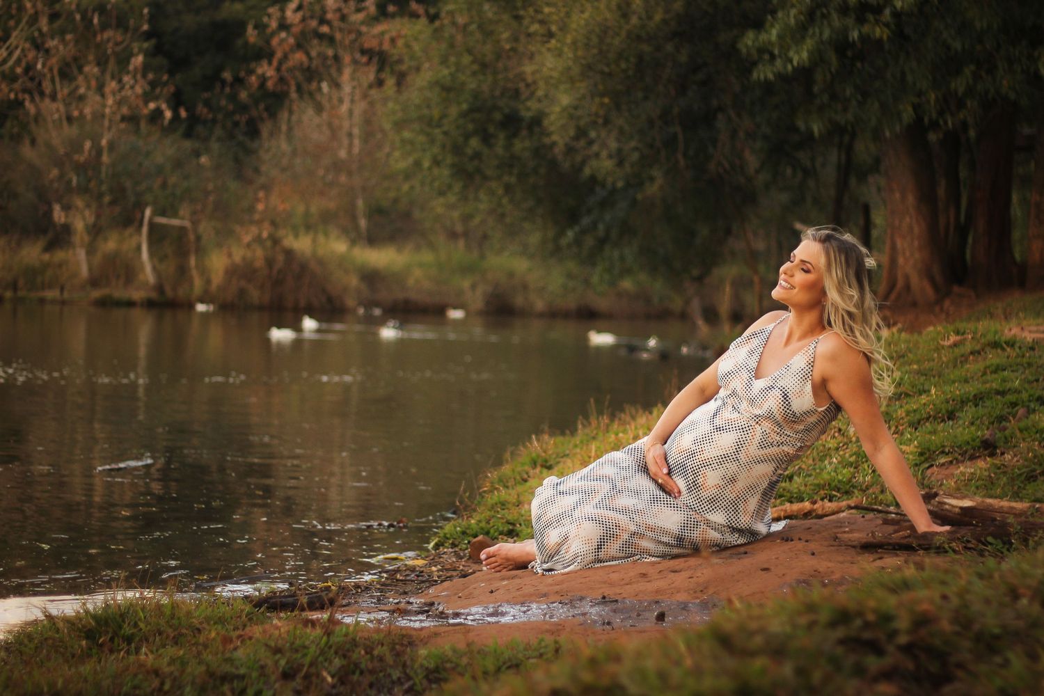 Woman in flowing dress reclining by a serene pond at sunset.