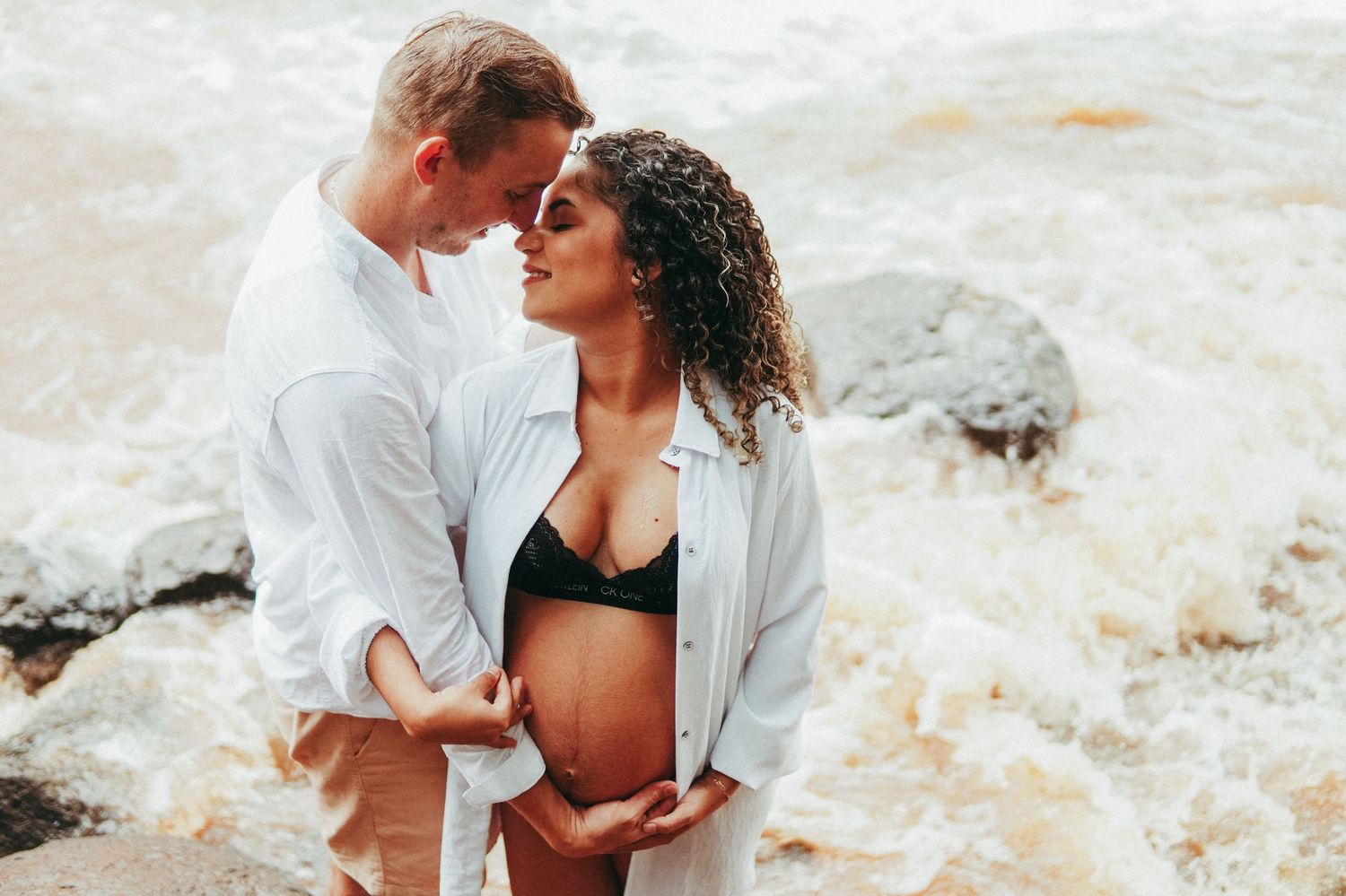 Romantic beach maternity photo with couple embracing against ocean waves.