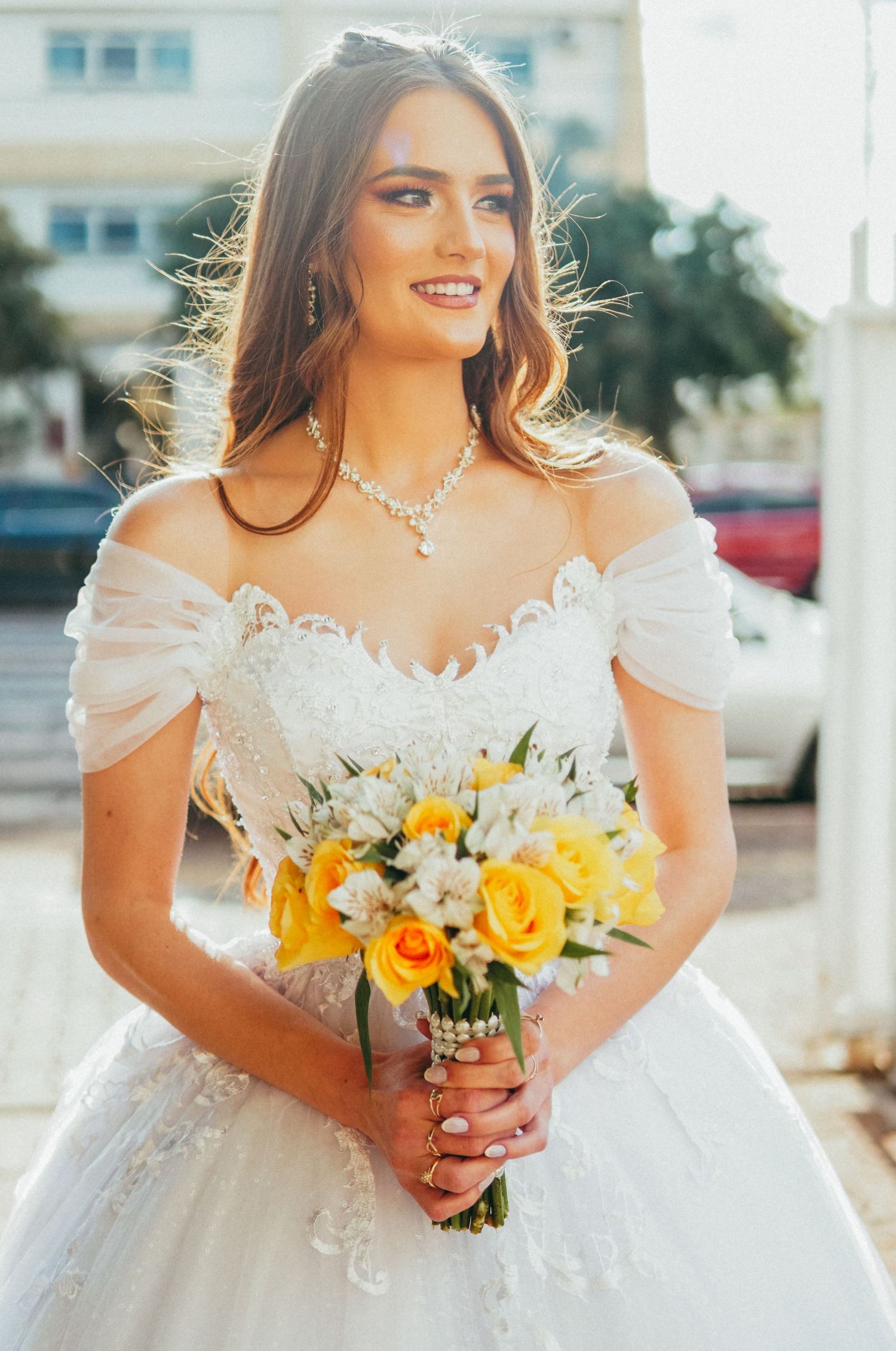 A radiant bride in an off-shoulder white gown holds a yellow and white bouquet in golden sunlight.