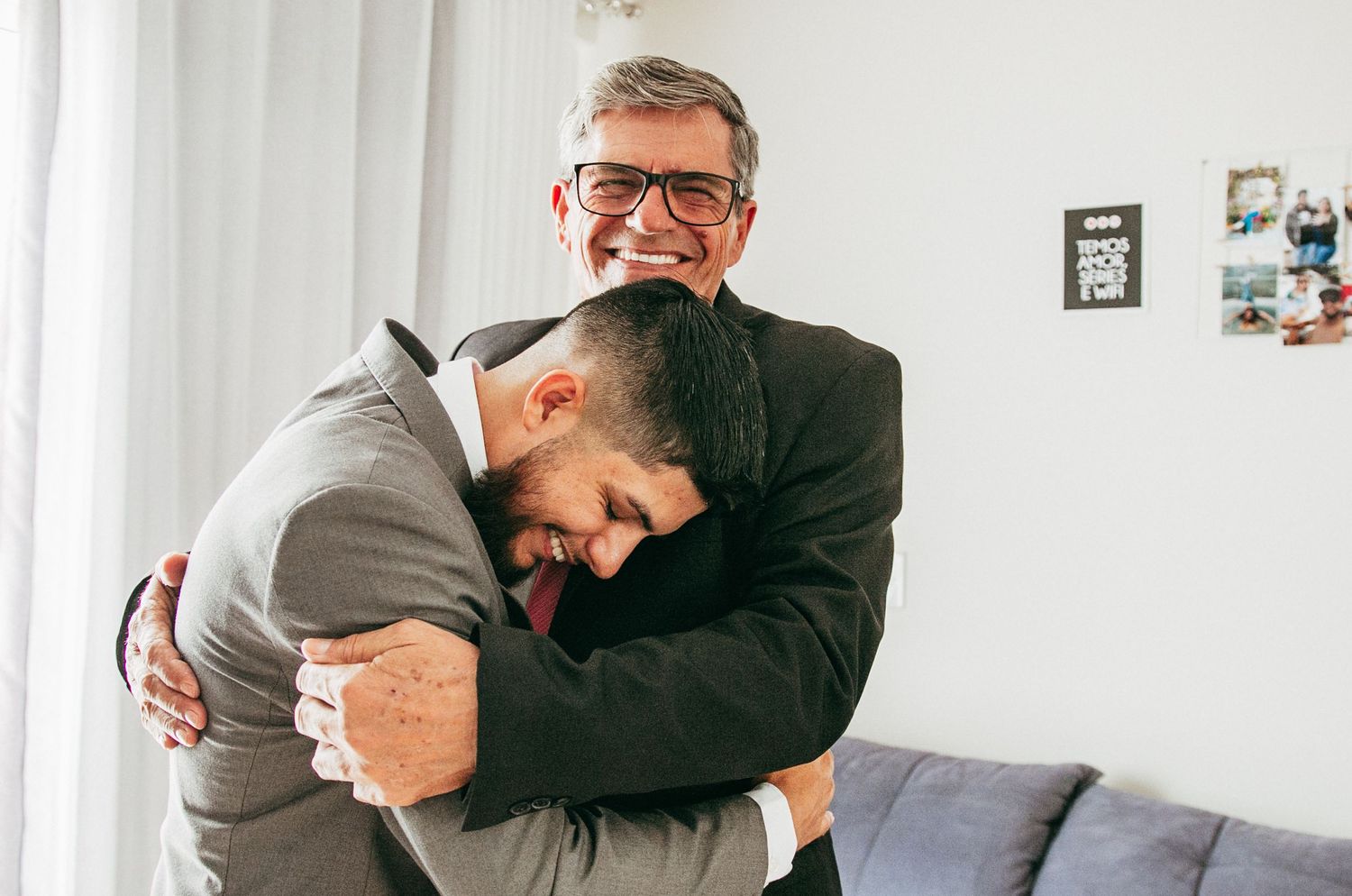 Two people share an emotional embrace while sitting on a gray couch in a bright living room.