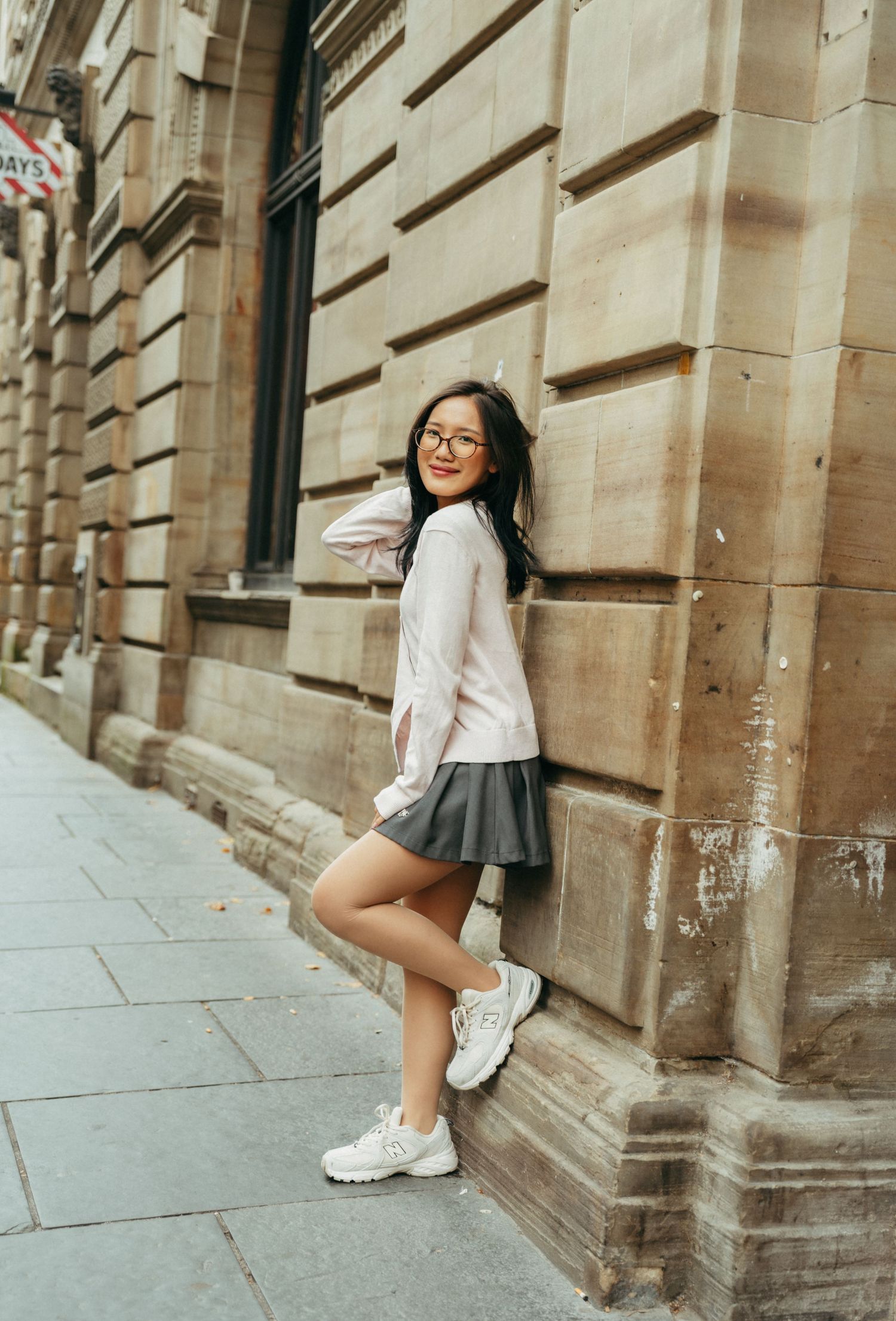 Young woman in gray mini skirt and white sweater leaning against a stone building wall on a city sidewalk.