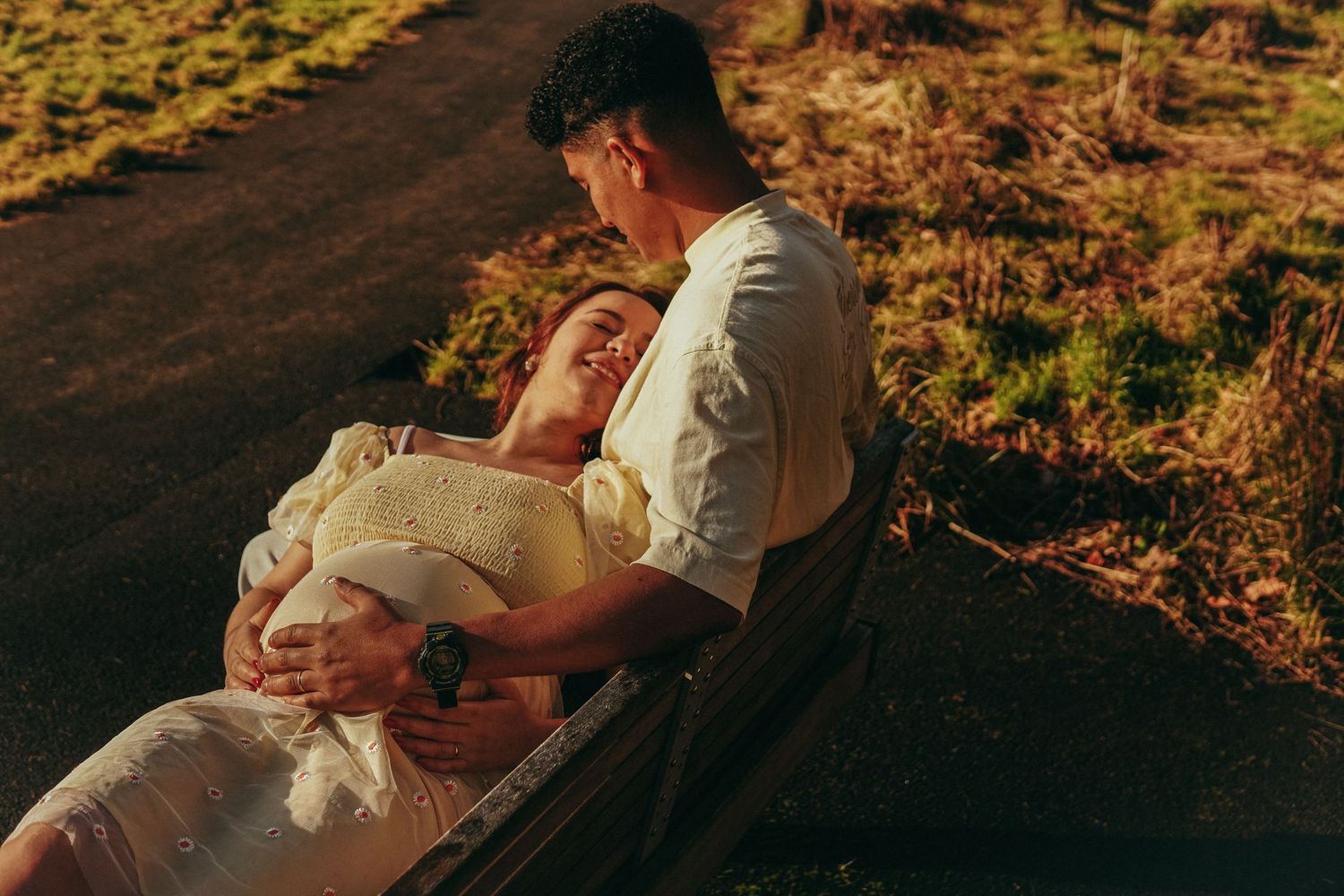 Couple sitting outdoors in golden light, woman reclining on man's lap, both cradling her pregnant belly.
