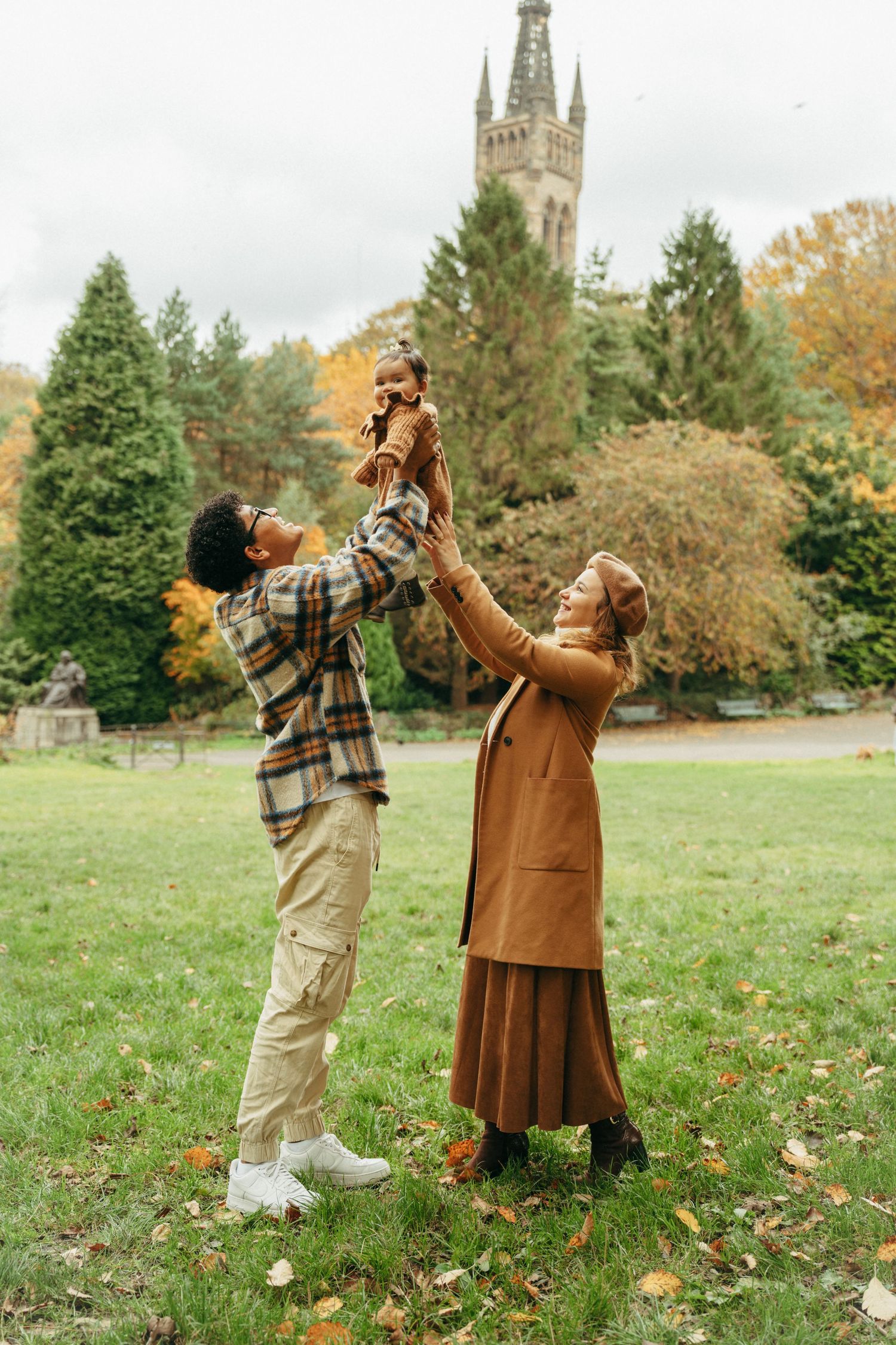 A joyful family lifting a toddler in an autumn park with trees and a historic tower in the background.