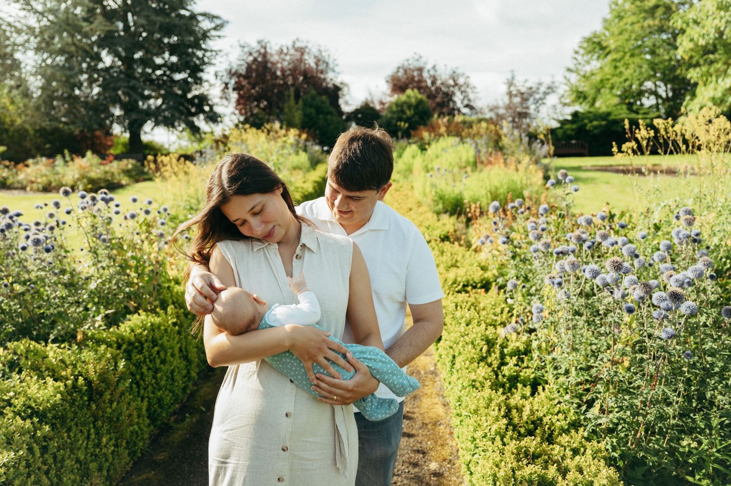A couple admires their newborn baby while walking through a lush garden with colorful wildflowers.