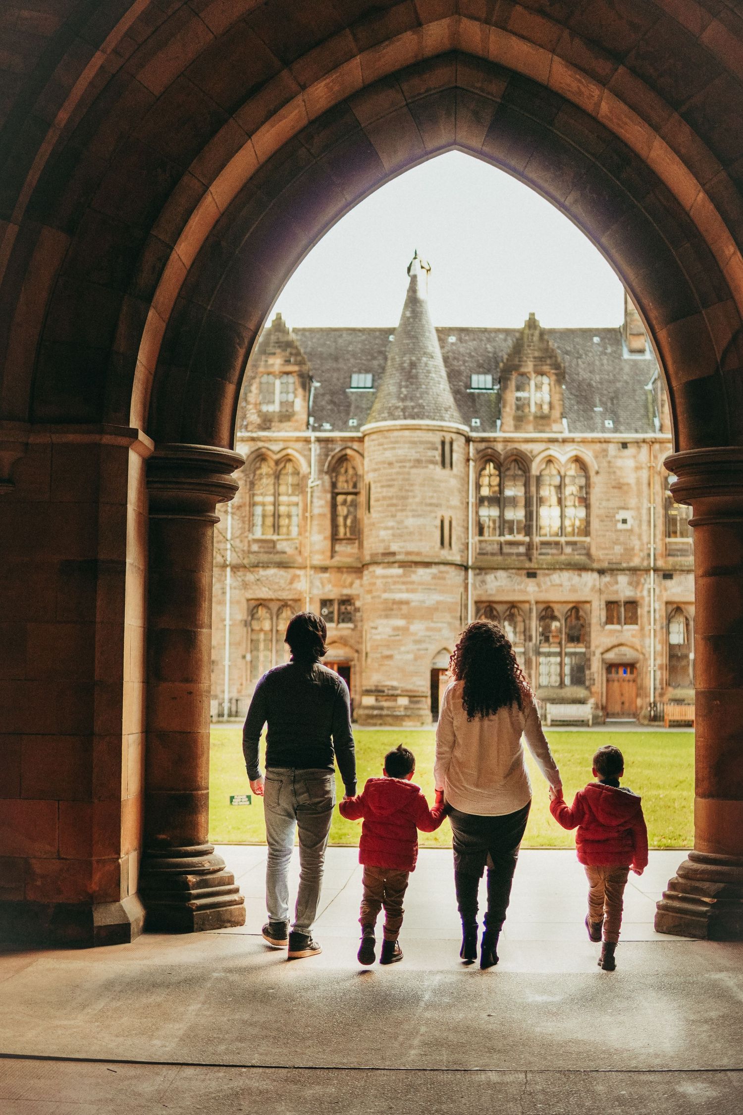 Family of four walks hand-in-hand through a Gothic stone archway toward a historic castle-like building.