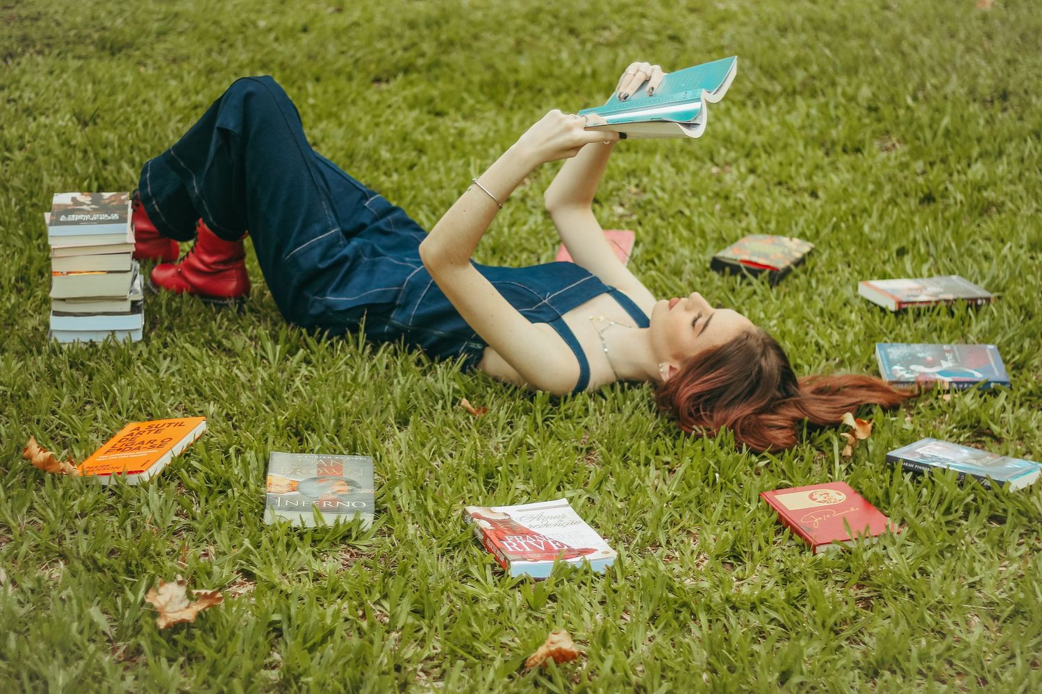 Person lying on grass reading a book surrounded by scattered books and colorful literature on a summer day.