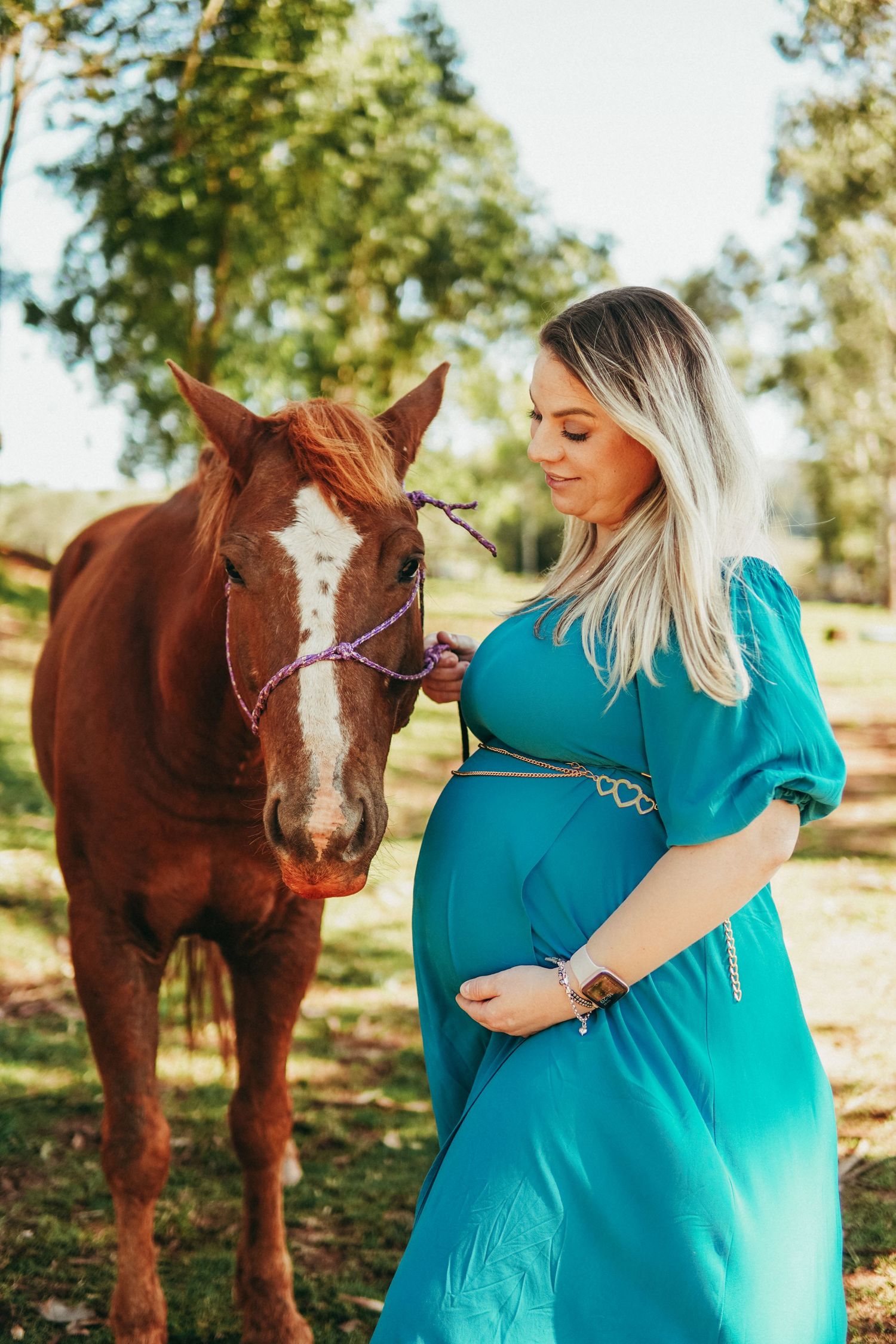 Pregnant woman in turquoise dress poses with brown horse in sunlit outdoor setting.