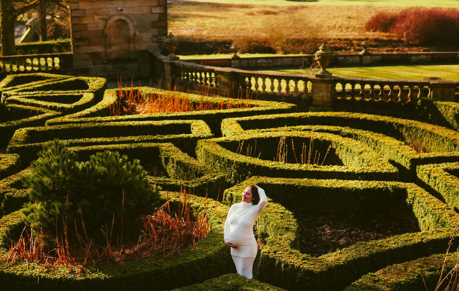 Person in white dress poses in an ornate formal garden with geometric hedgerows and stone balustrade.
