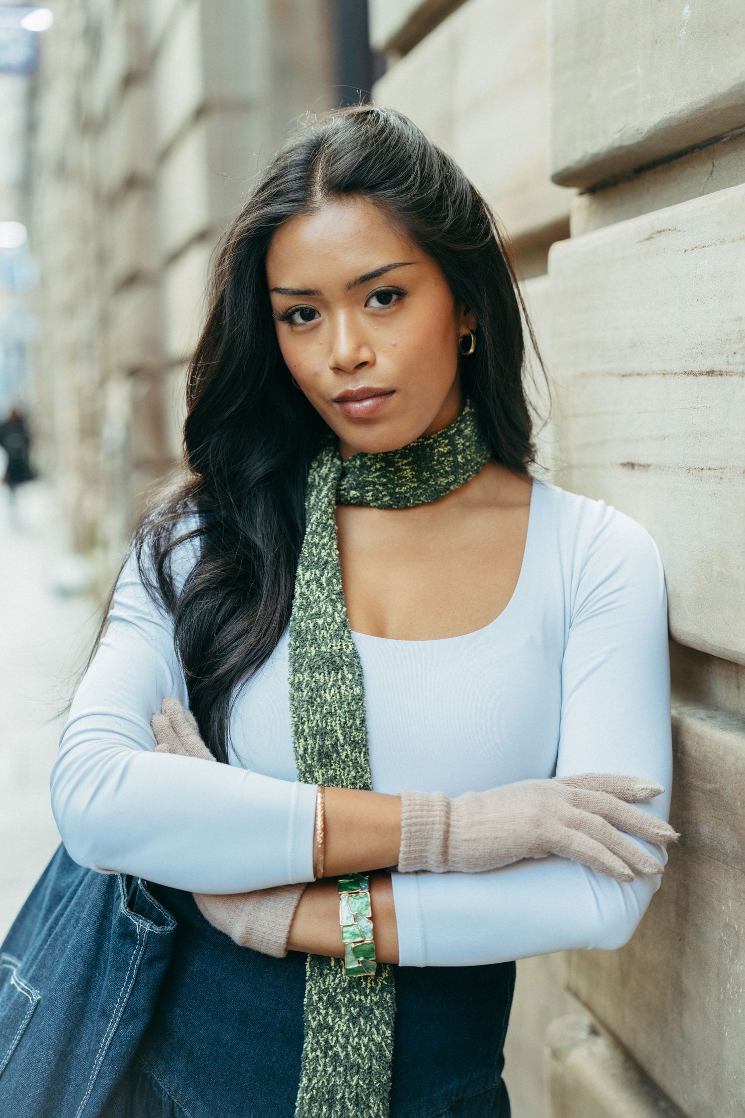 Young woman with long dark hair wearing a white top and green scarf, arms crossed, leaning against a stone wall outdoors.