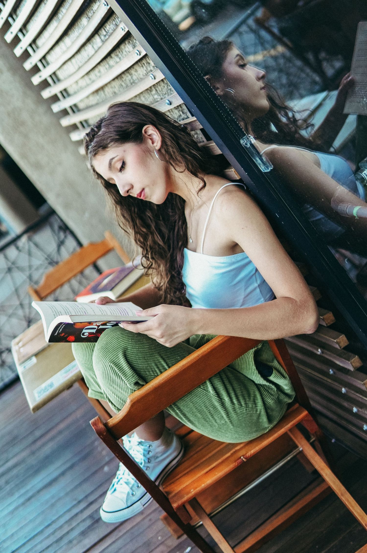 A person in a blue tank top and green pants reads while sitting in a wooden folding chair by a window.