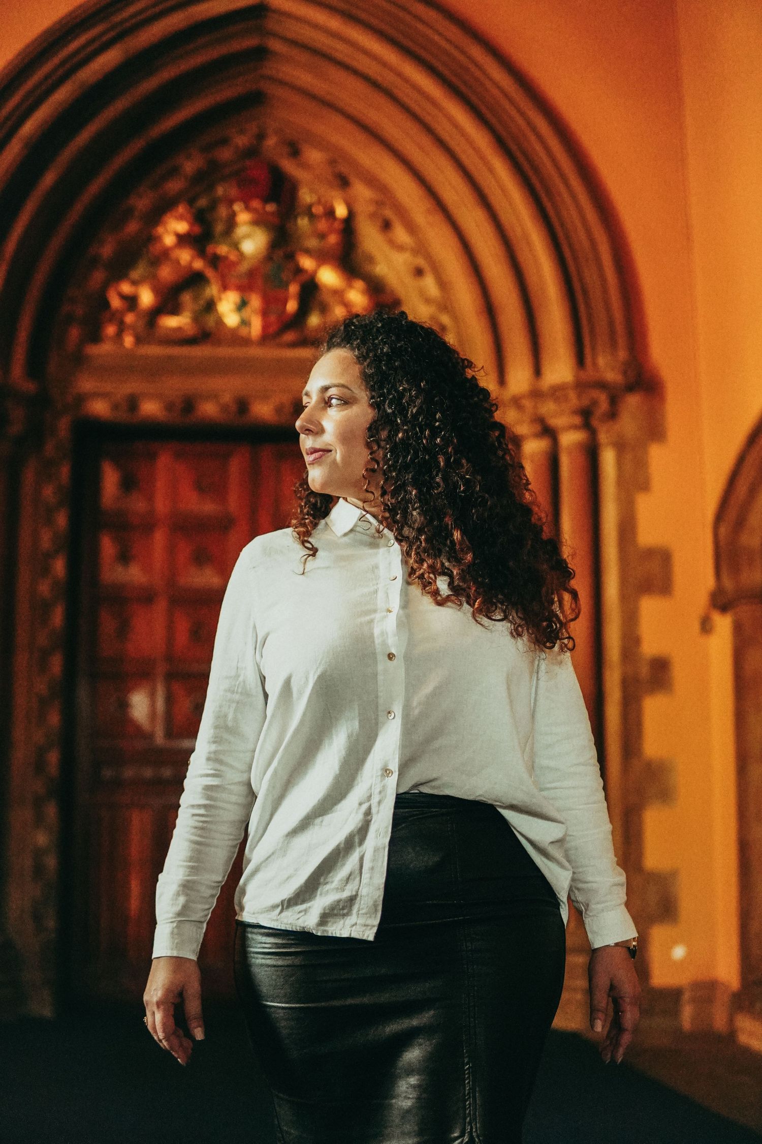 A person with curly hair wearing a white button-up shirt and black leather skirt stands in front of an ornate archway.