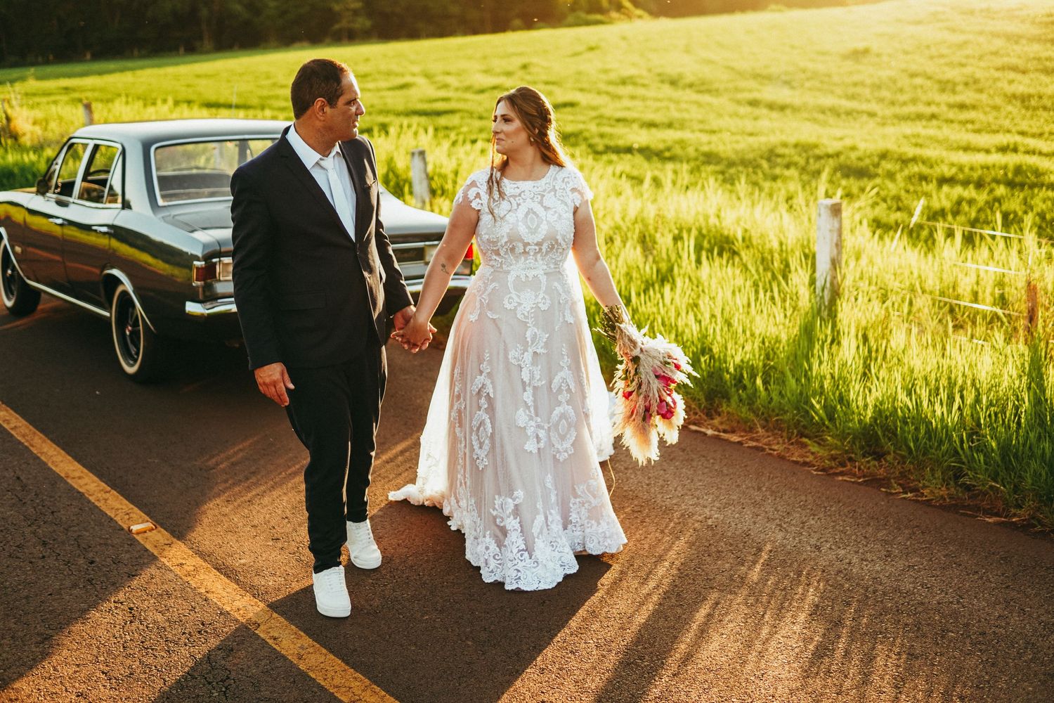 A couple holding hands on a country road at sunset with a vintage black car and golden sunlit field in background.