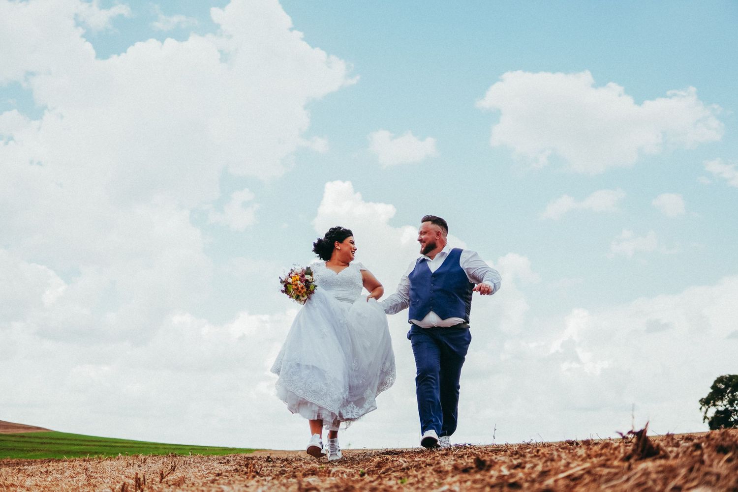 A happy bride and groom joyfully run across a hilltop together against a bright blue cloudy sky.
