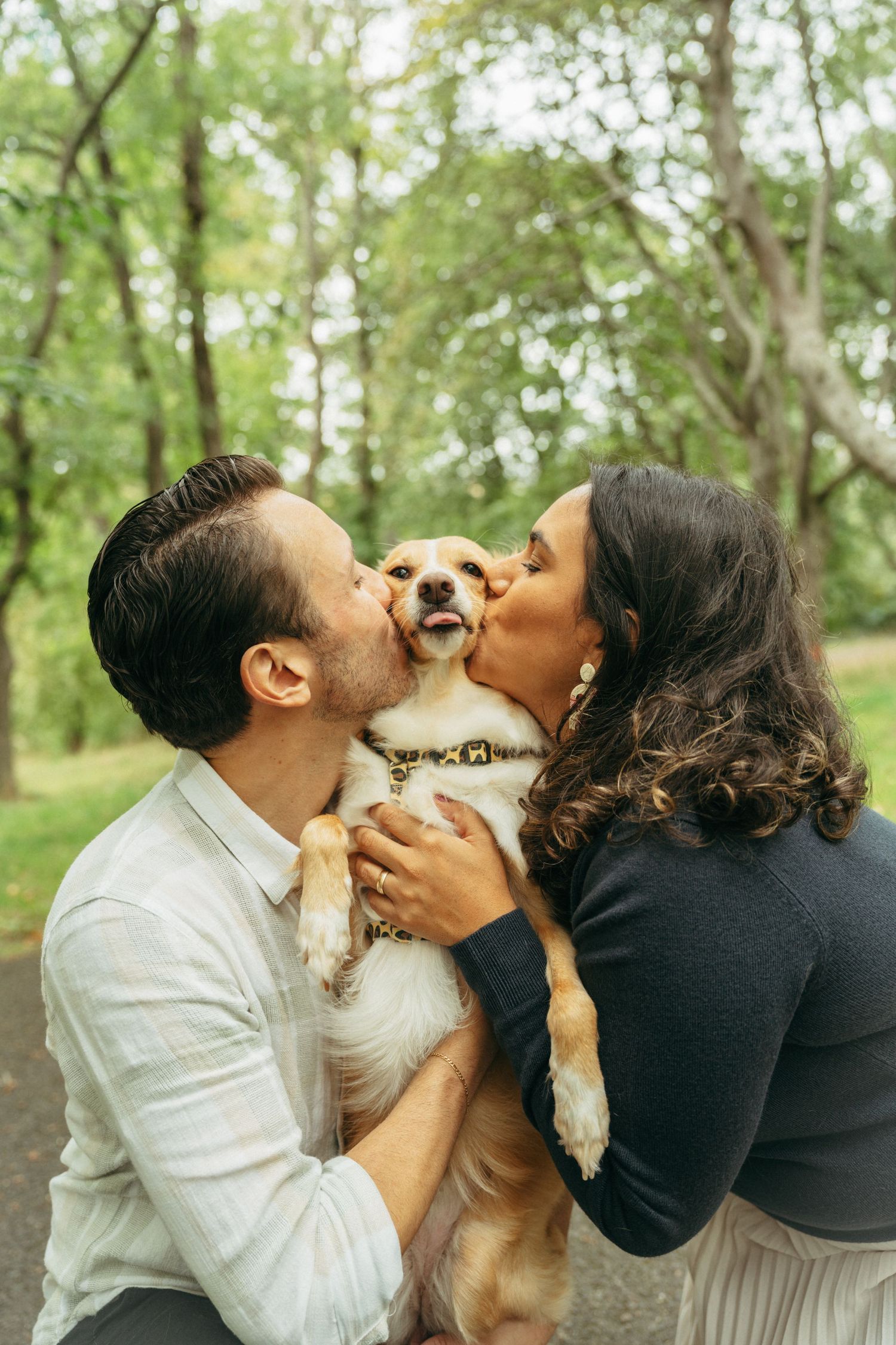 Couple kissing their golden retriever dog on both cheeks in a lush green park setting.