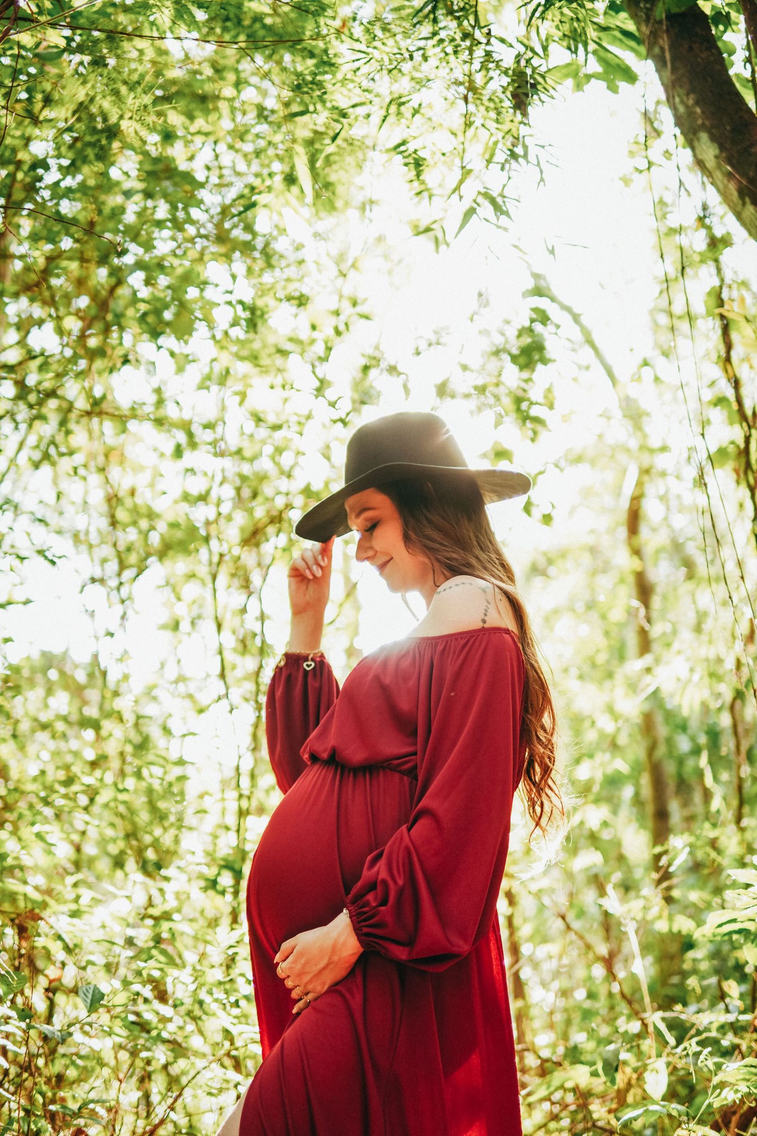 Ethereal maternity portrait of woman in red dress and hat standing among sun-dappled trees.