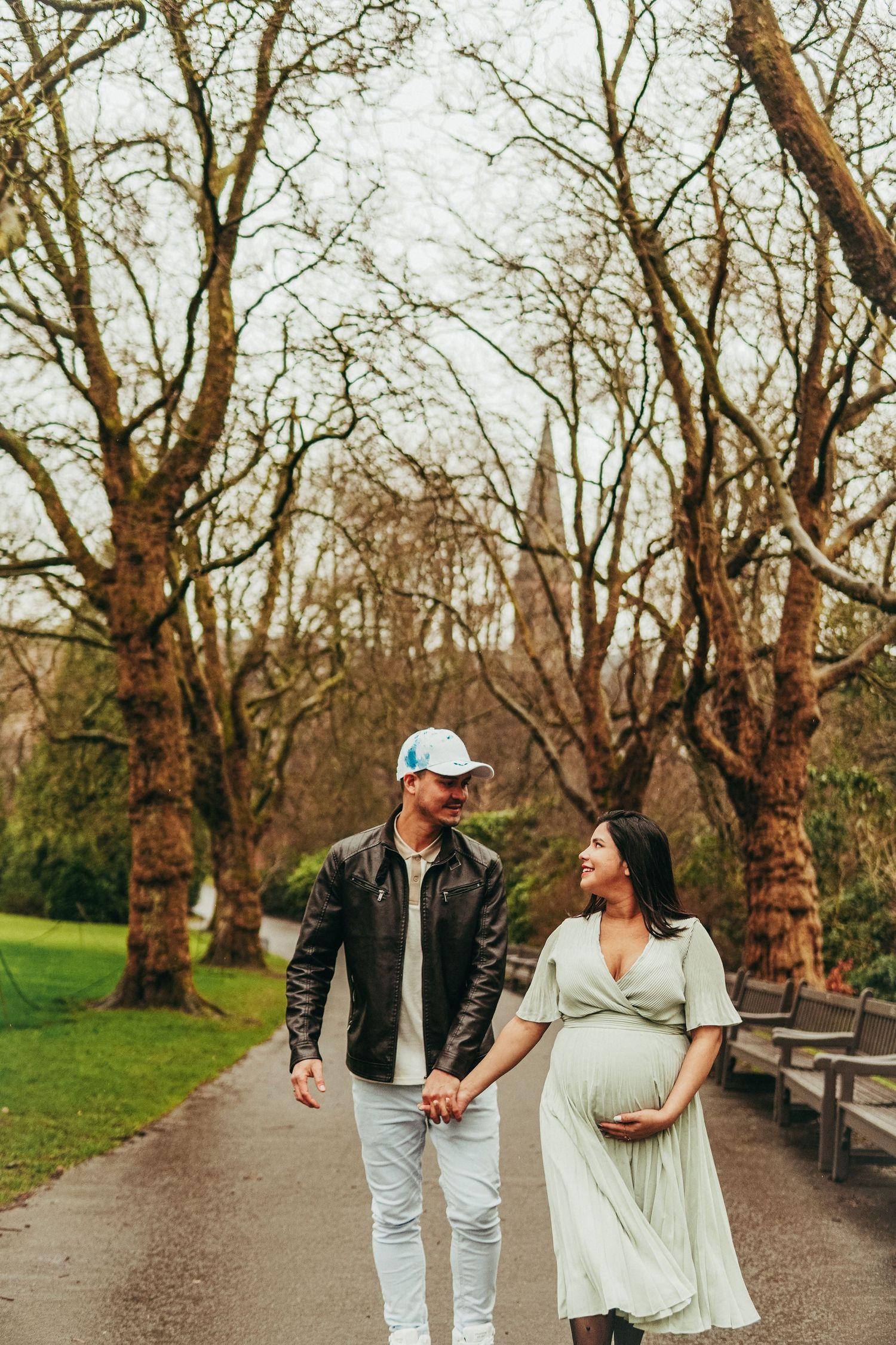 Pregnant couple holding hands walking on park path, man in leather jacket and cap, woman in sage green maternity dress.