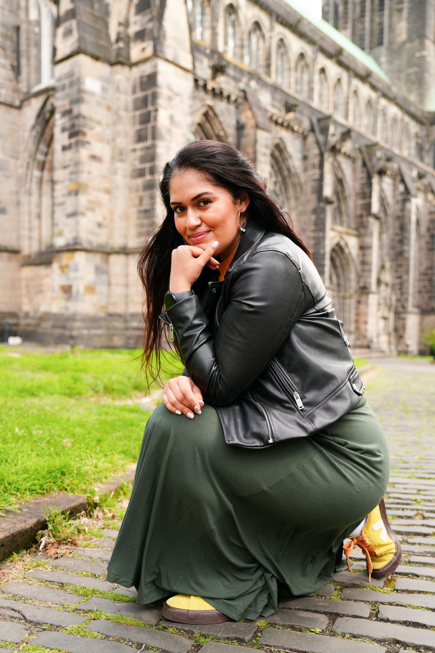 A young woman in a leather jacket and green skirt crouches on cobblestones outside a historic stone cathedral.
