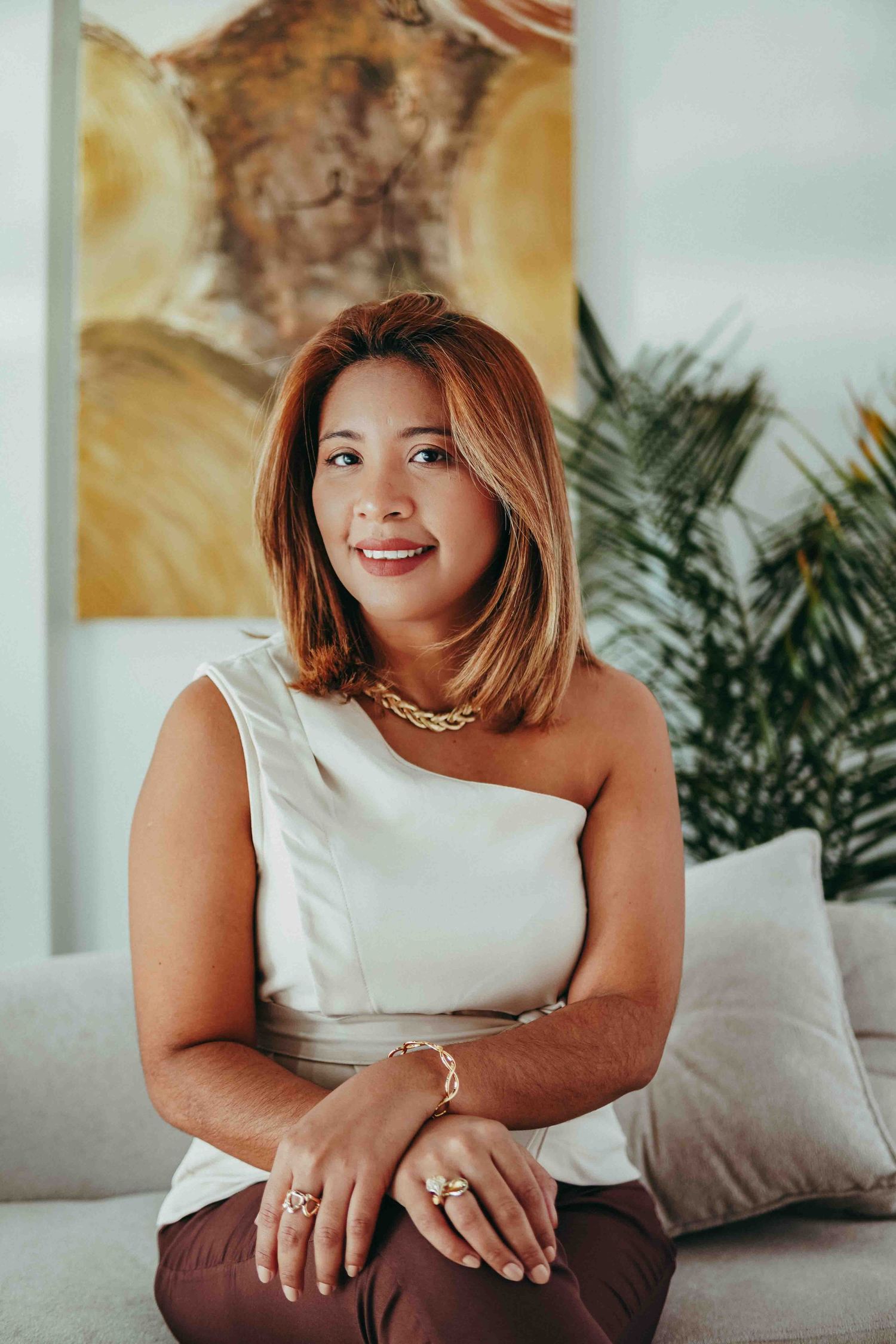 A person in a white one-shoulder top sits on a couch with palm plants and wall art visible in the background.