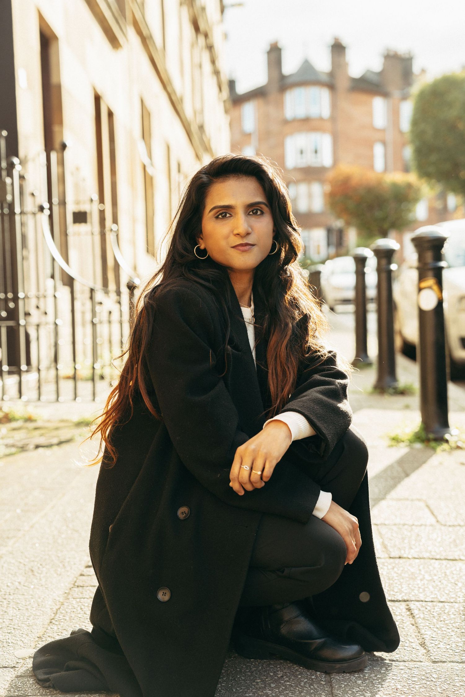 Young woman in black coat crouching on a sunlit city sidewalk, posing confidently with long dark hair.