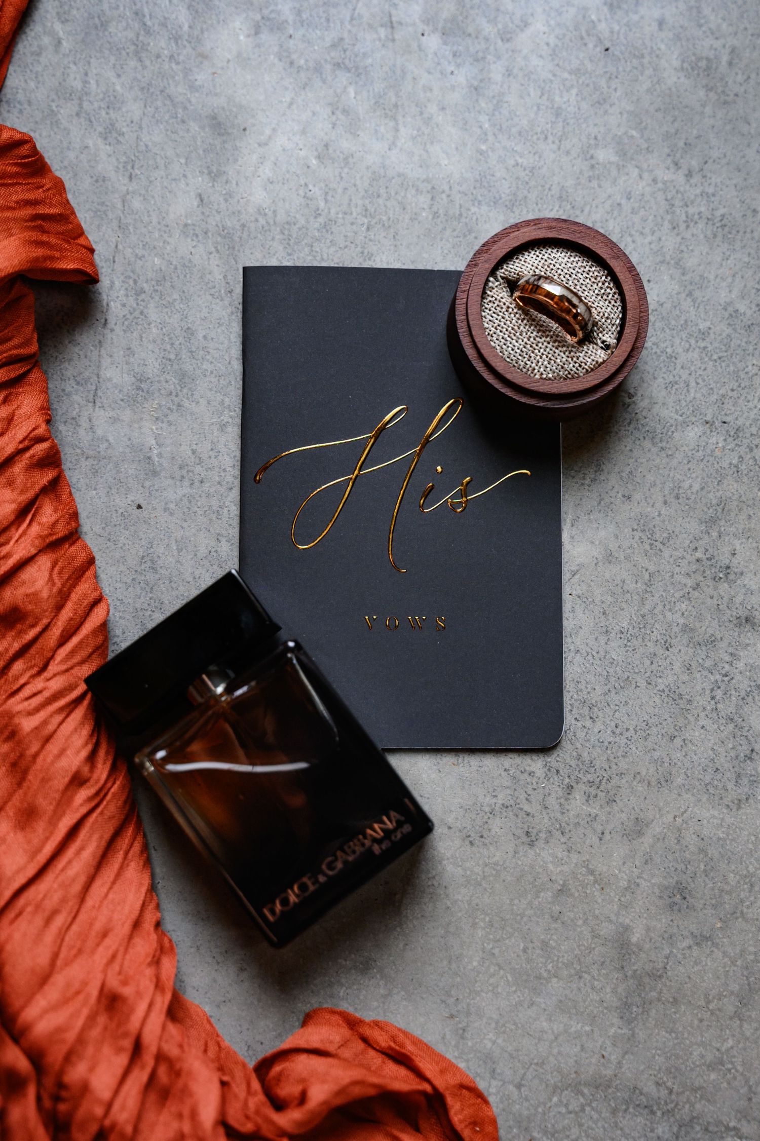 Black wedding vows book with gold lettering alongside perfume bottle and ring box on gray surface with orange fabric.