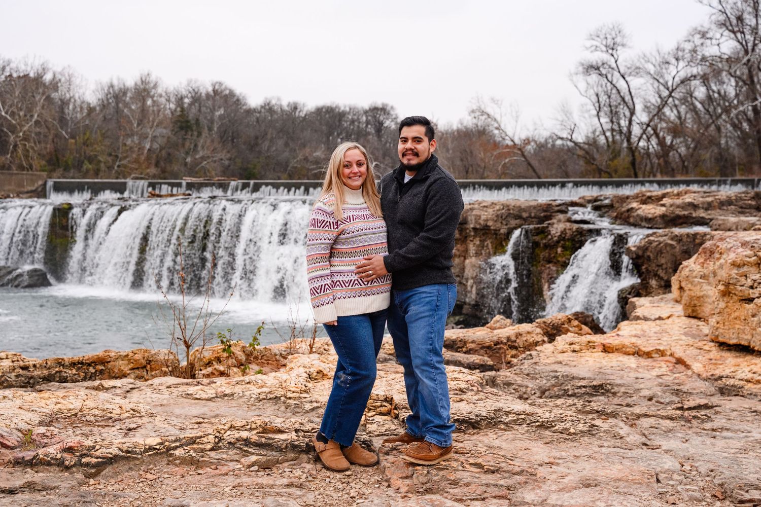 A couple stands together near a scenic waterfall at Joplin's Grand Falls in Missouri during winter.
