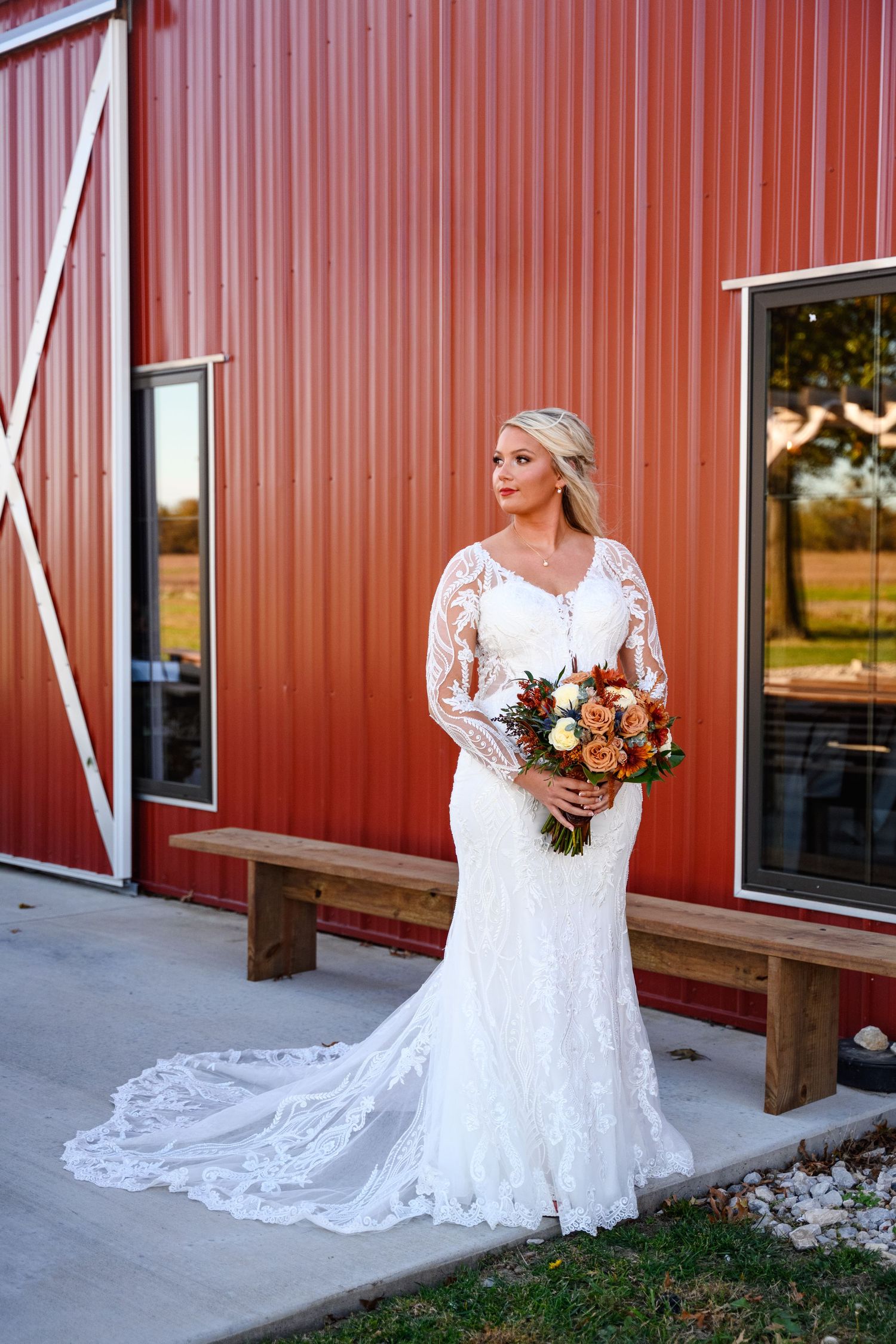 A bride in a lace wedding dress poses against a rustic red barn in a series of bridal portraits at sunset.