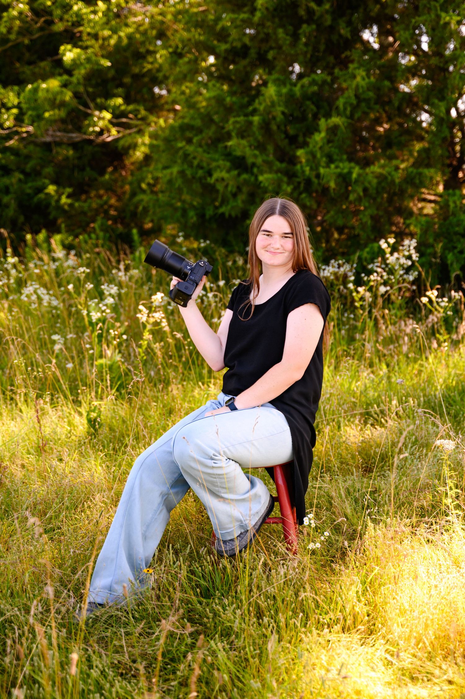 Amber Ross- photographer with camera sitting in golden sunlit field with green trees in background.
