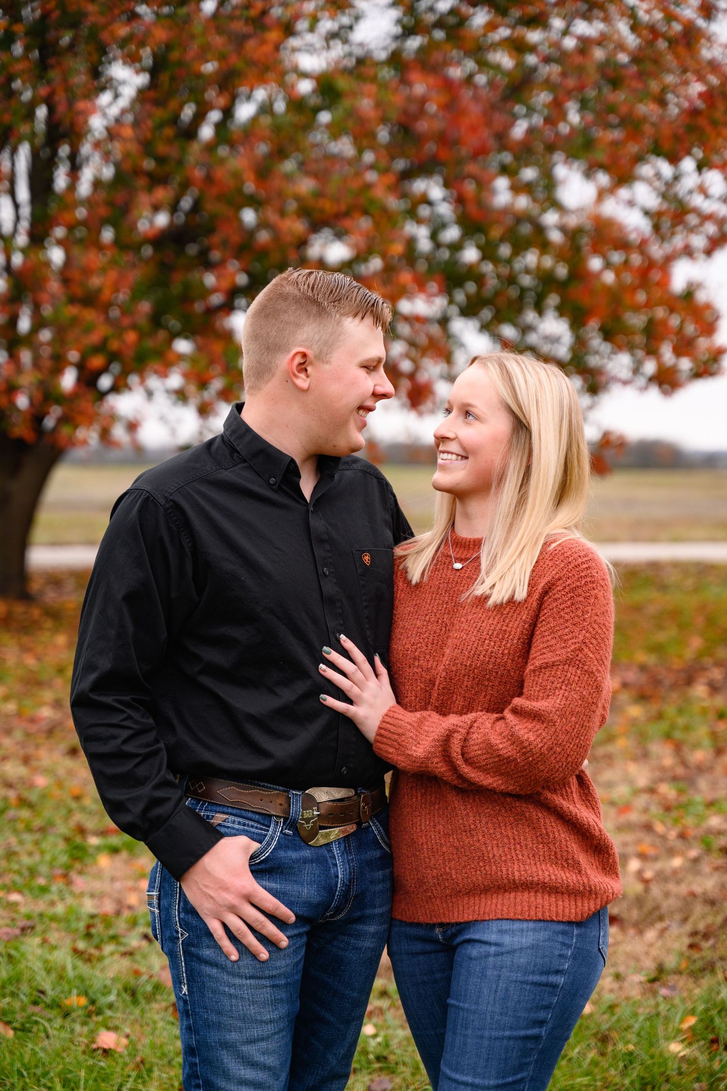 A couple in casual fall attire embraces outdoors against a backdrop of autumn foliage and fallen leaves.