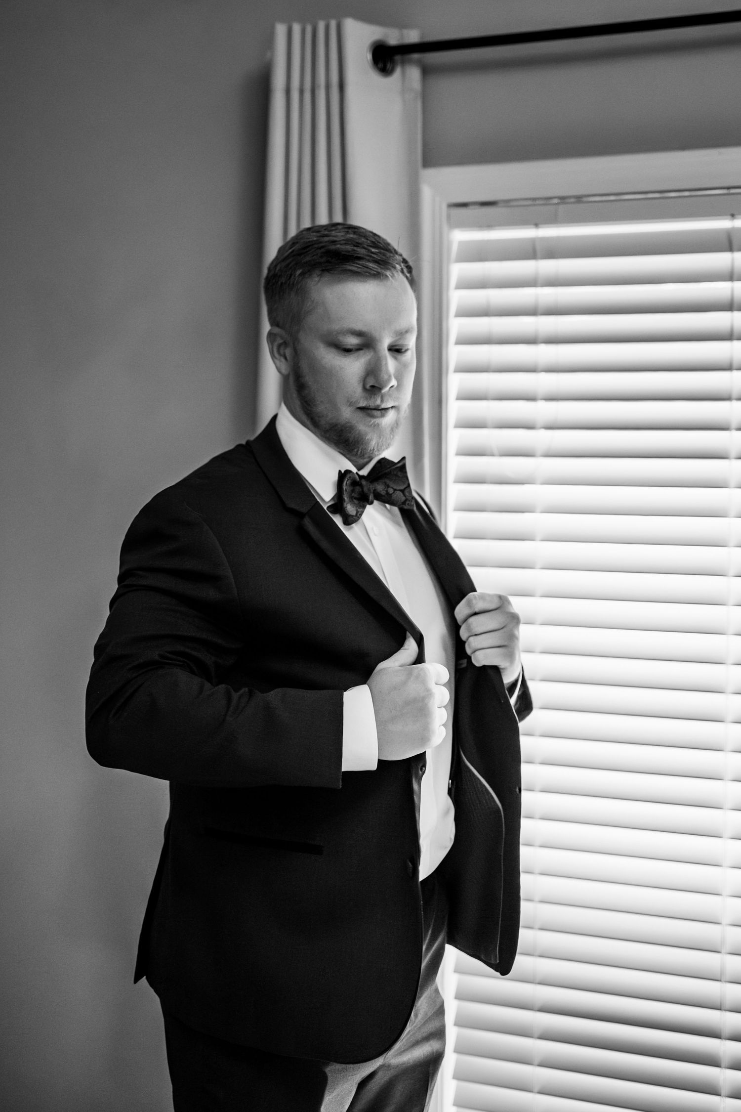 A groom adjusting his jacket near a window, while getting ready for his wedding in Arkansas.