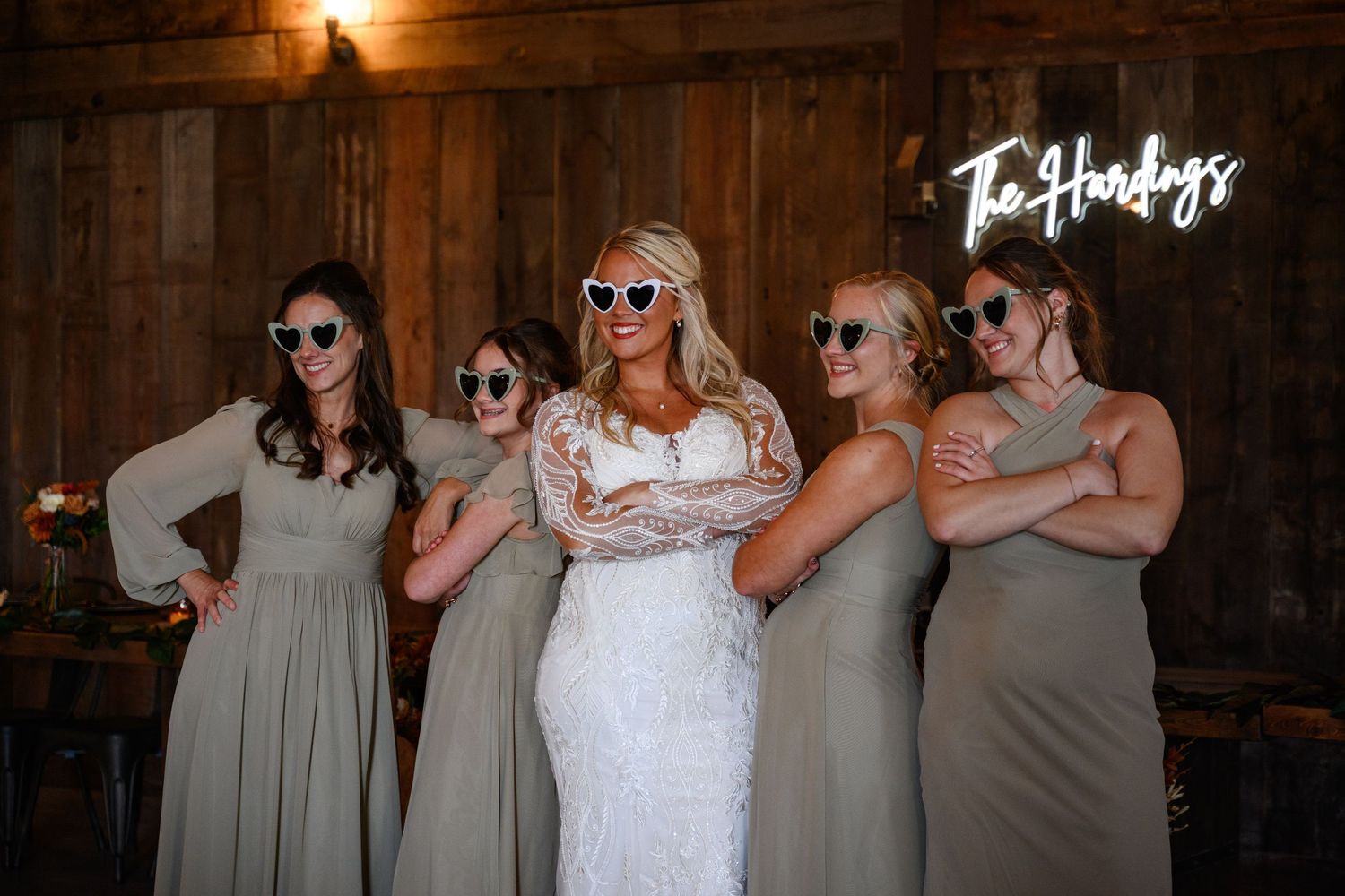 A wedding party in matching outfits poses playfully with sunglasses against a rustic wood wall at Camp 400.
