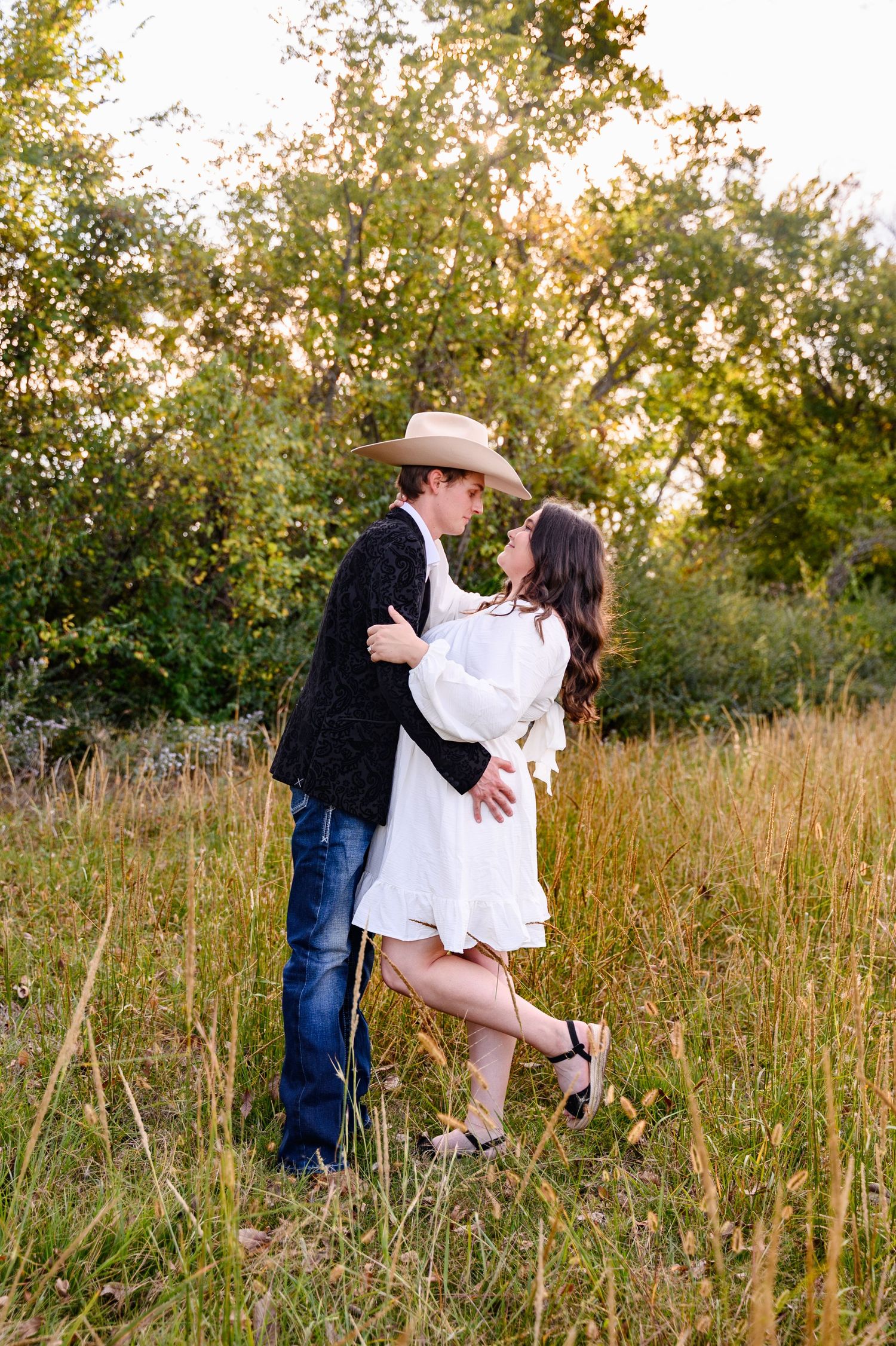 A couple shares romantic kisses in a sunlit meadow with tall grass and trees during golden hour.