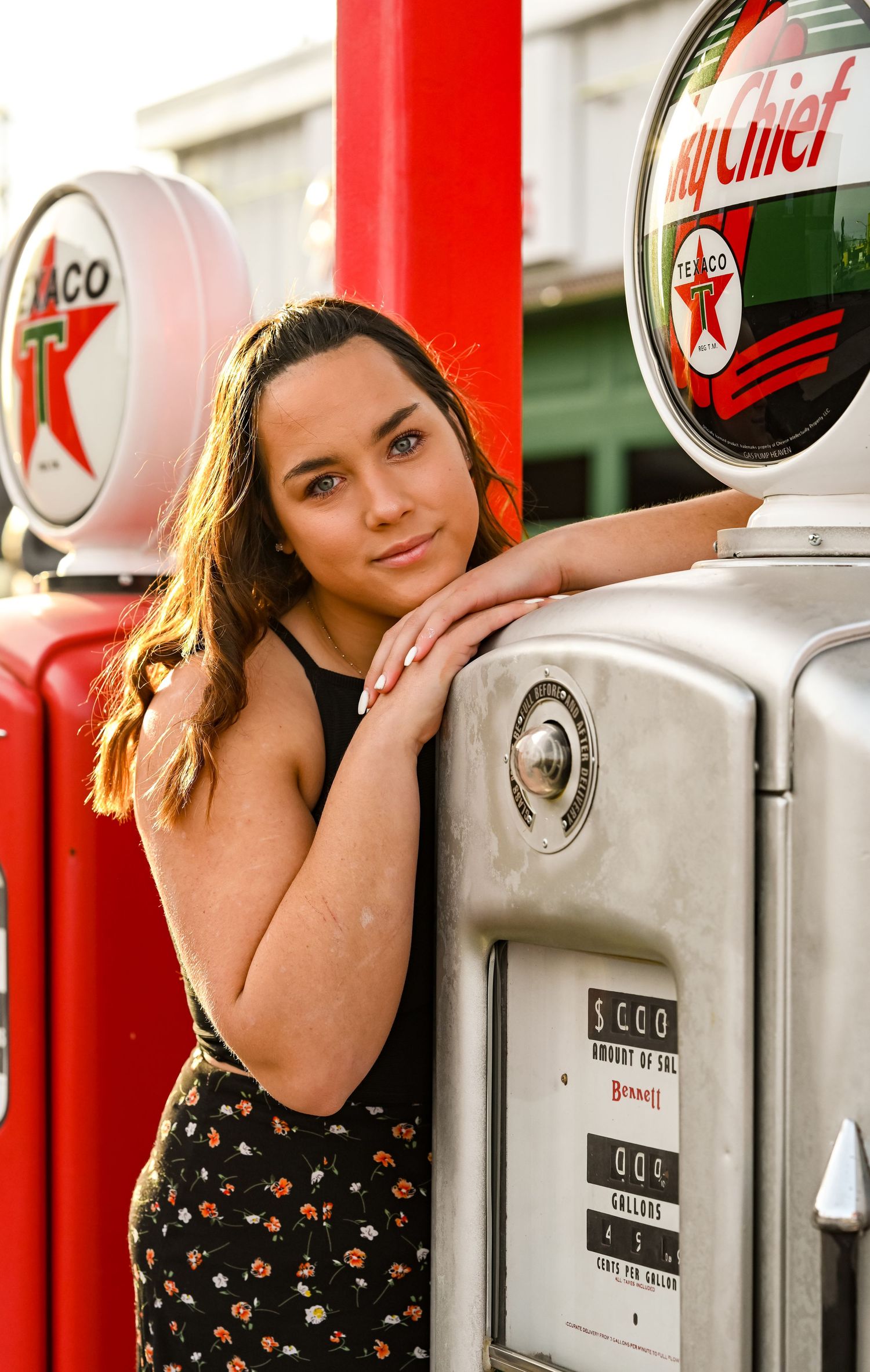 A senior leaning on a vintage gas pump, with the sunlight glowing behind her.