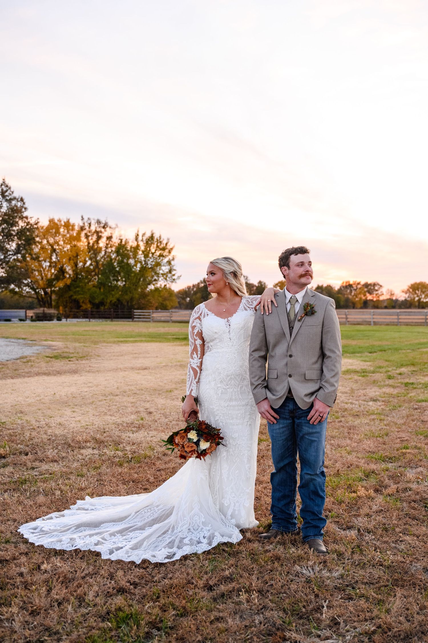 A couple poses together in a rustic outdoor setting at sunset with a long white wedding dress trailing in the grass.