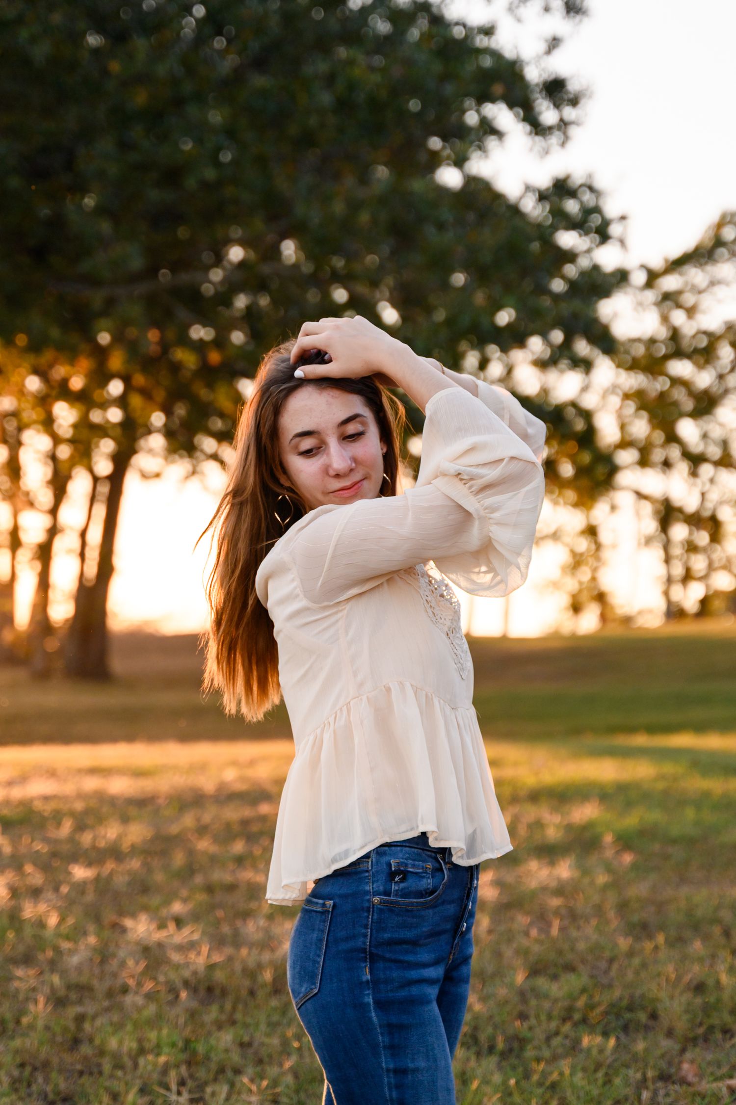 A senior girl in a cream blouse and jeans poses outdoors during golden hour sunset with trees in the background.
