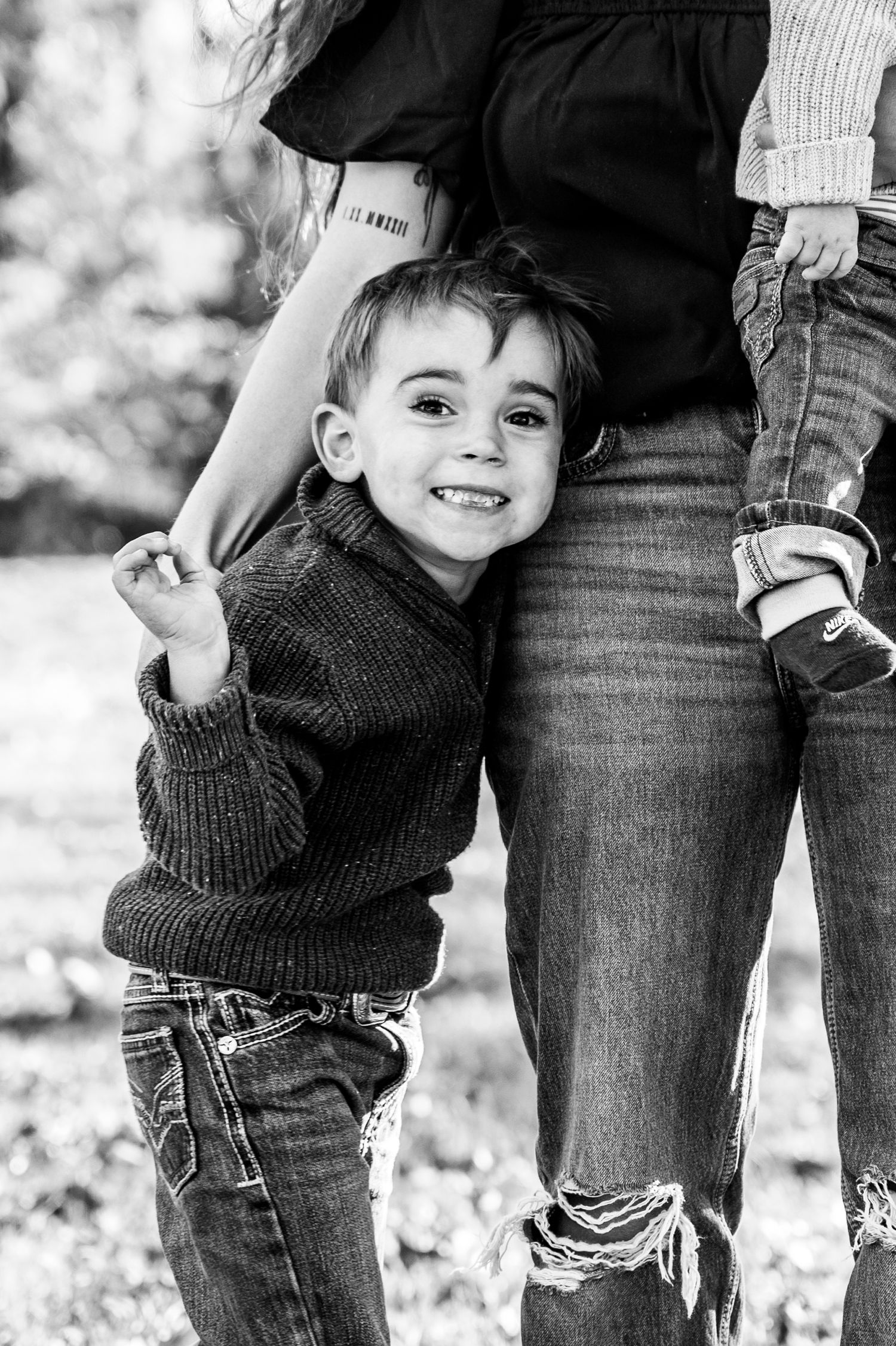 A young boy smiling at the camera, hugging his mom's leg.