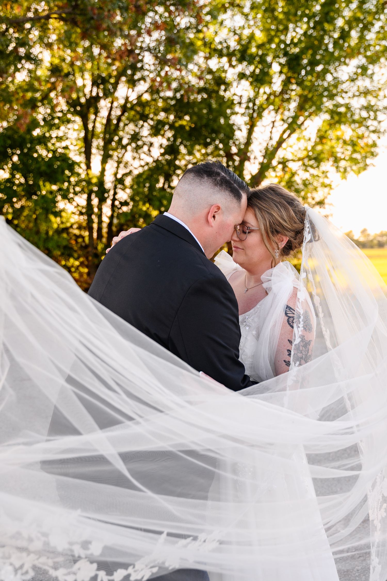 A wedding couple shares a kiss as the bride's long veil flows in the sunlight through trees.