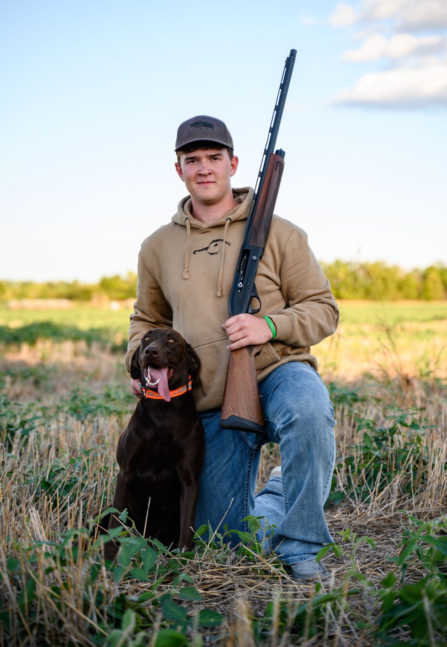 A senior and a chocolate Labrador retriever sit together in a field.